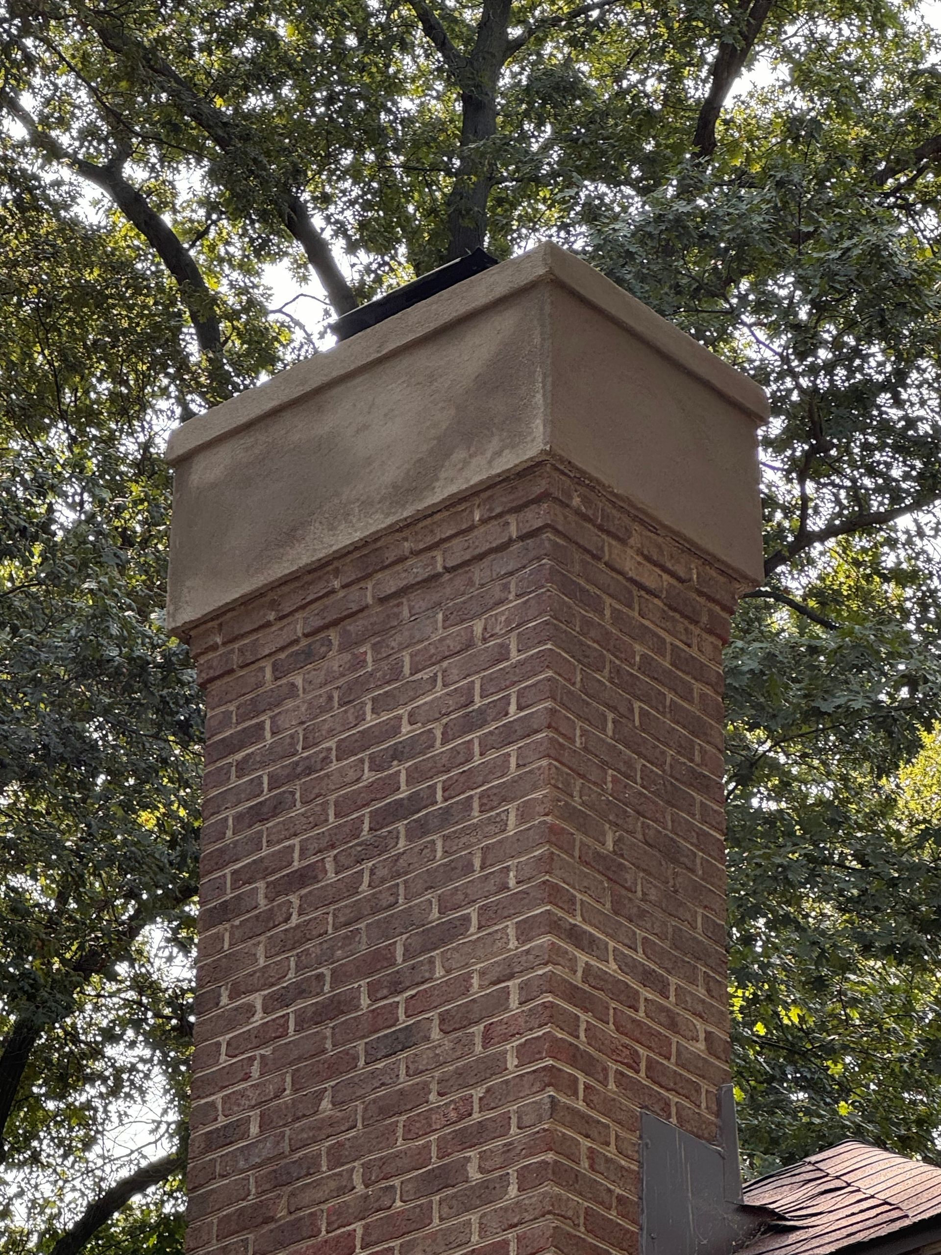 Tall brick chimney with a concrete cap against leafy trees and a tiled roof