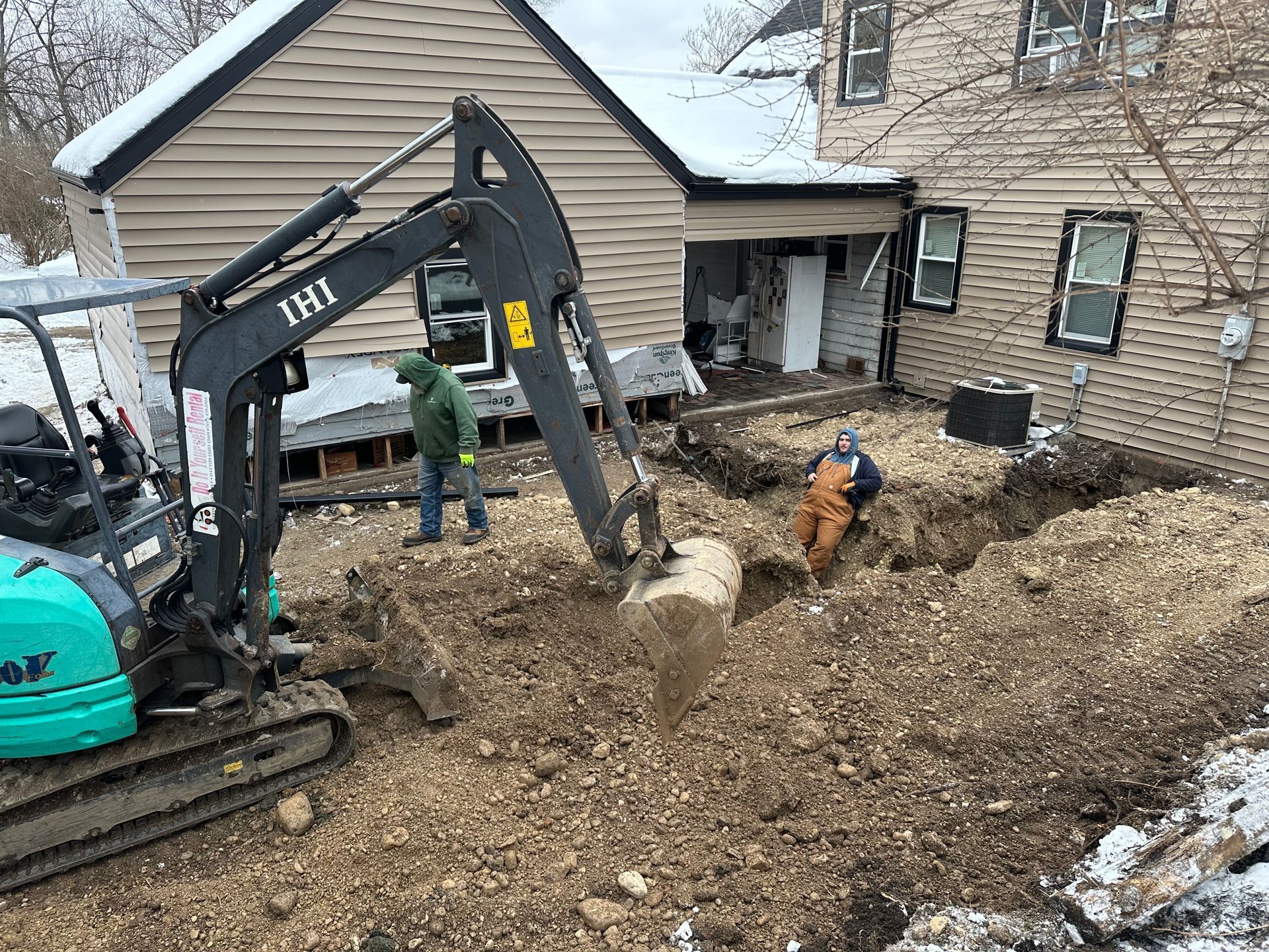 Excavator and workers digging a muddy residential yard beside a house and garage