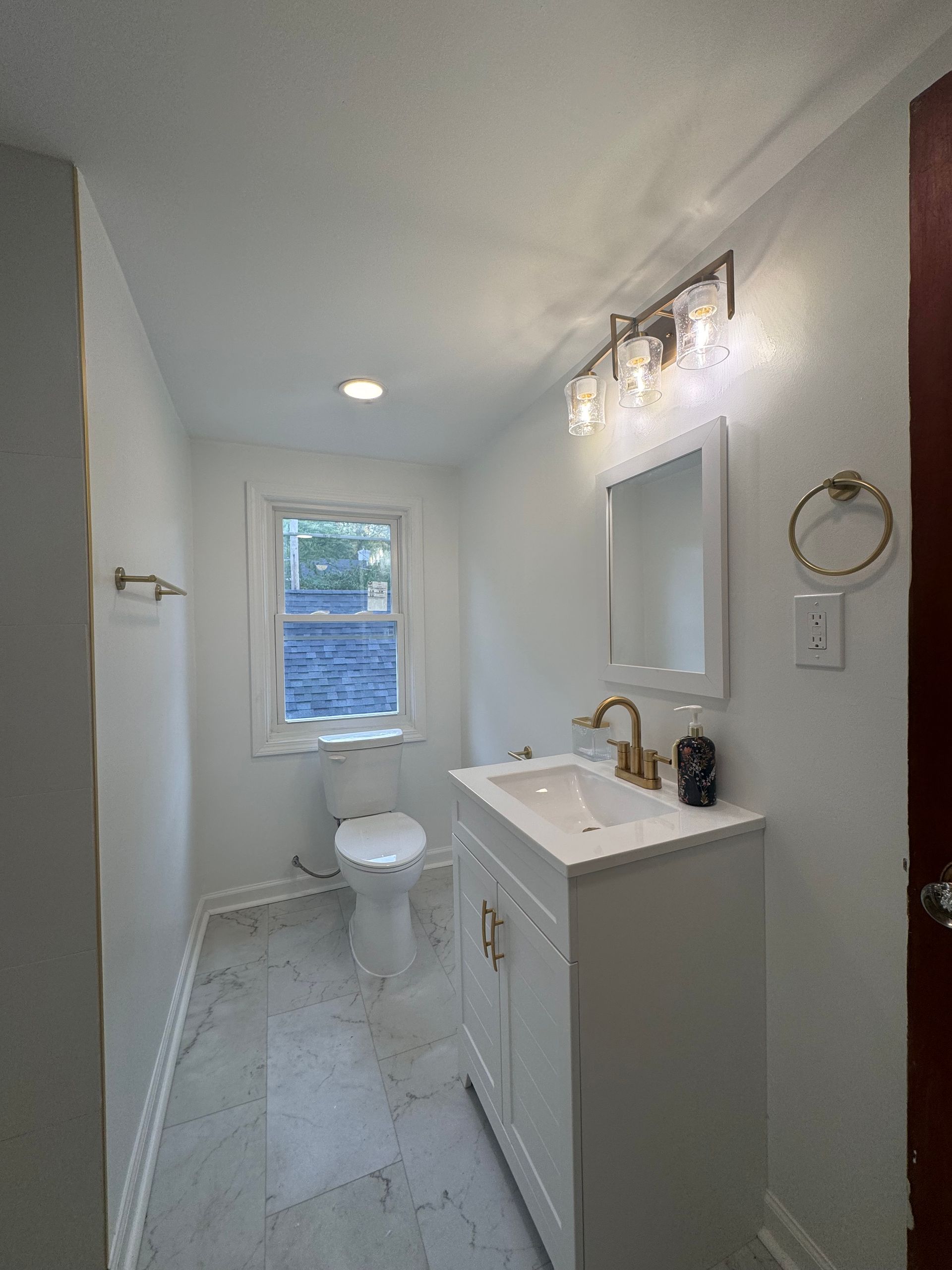Narrow white bathroom with sink vanity, toilet, window, and wall light.
