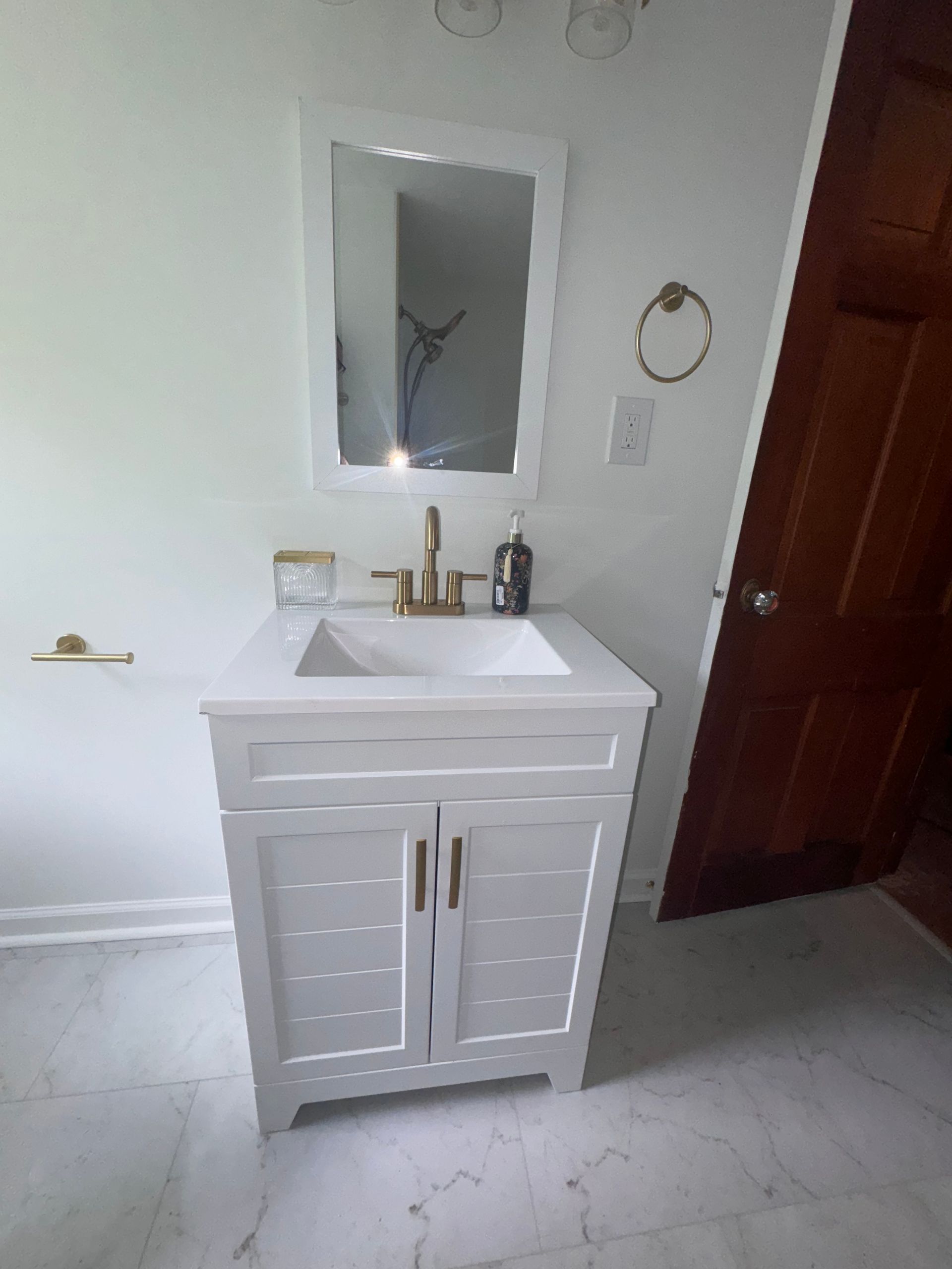 White bathroom vanity with sink, mirror, and brown door on a light tiled floor.