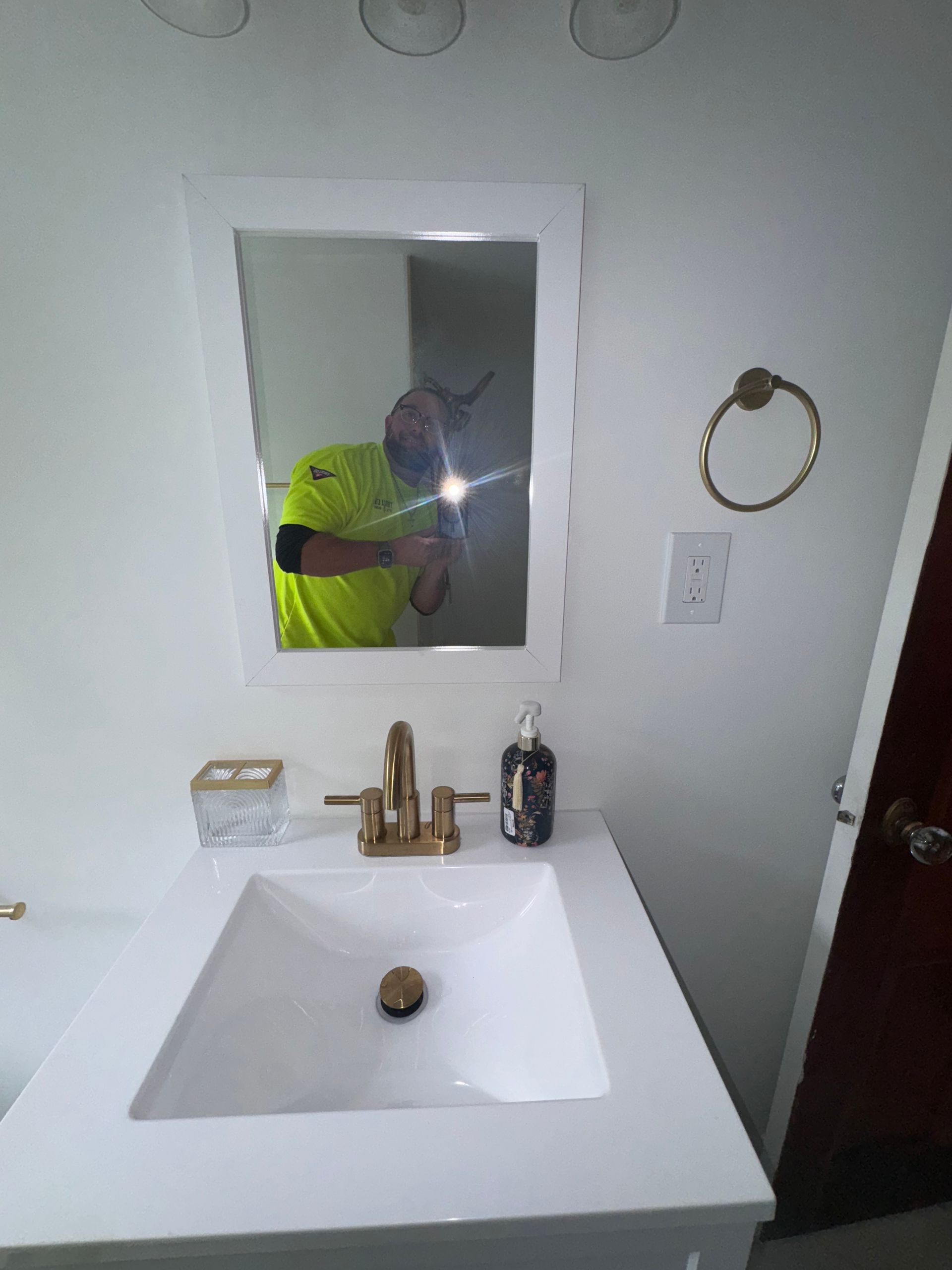 Bathroom sink with gold faucet, mirror, towel ring, and a person in a neon shirt reflected taking a photo.