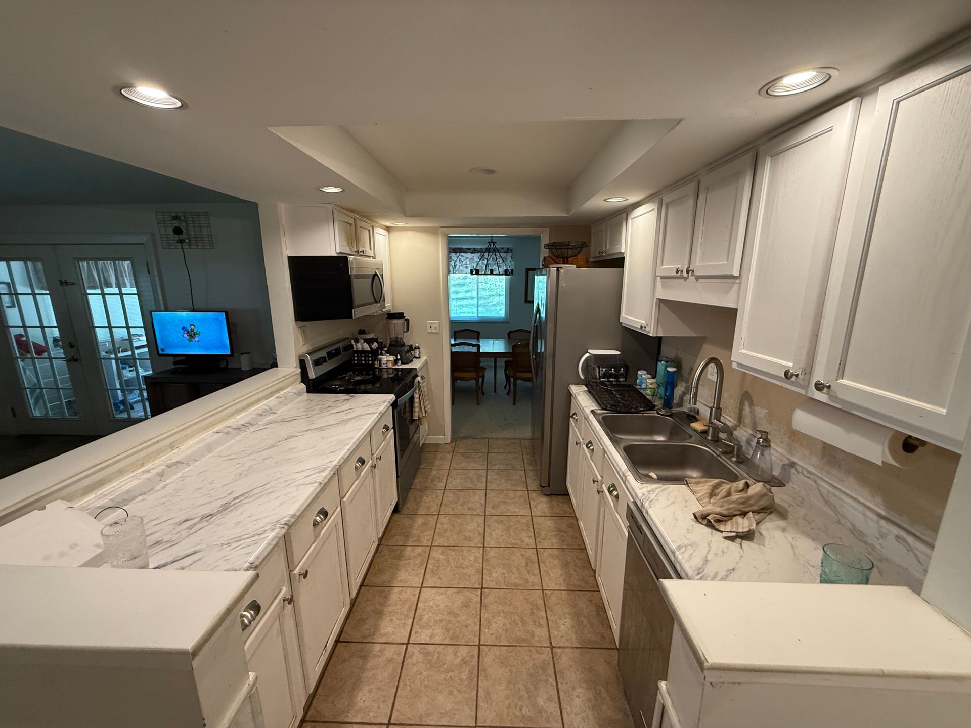 Bright kitchen with white cabinets, tiled floor, and a long galley layout leading to a dining area.