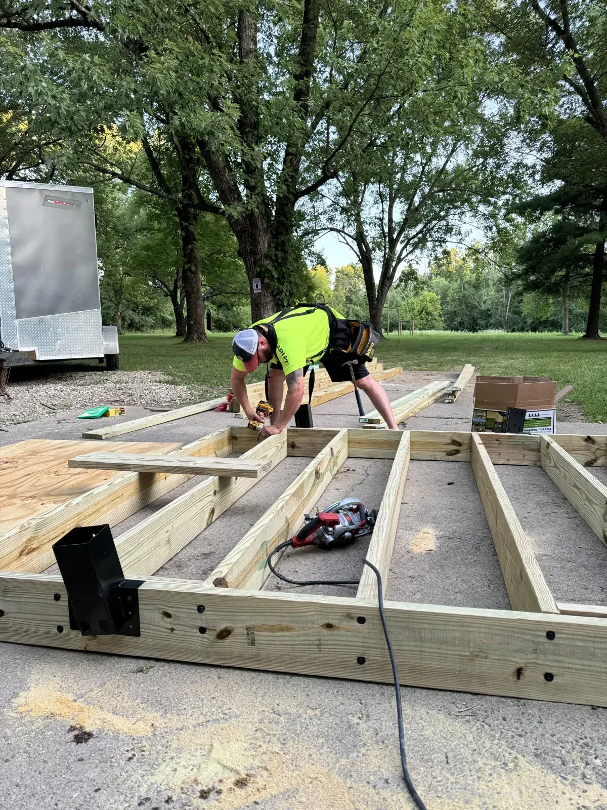 Workers assembling a wooden deck frame outdoors beside a trailer and trees