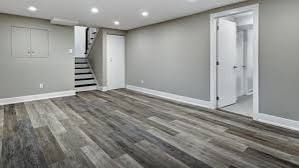 Empty finished basement with gray walls, wood-look flooring, recessed lights, stairs, and a doorway to a bathroom.