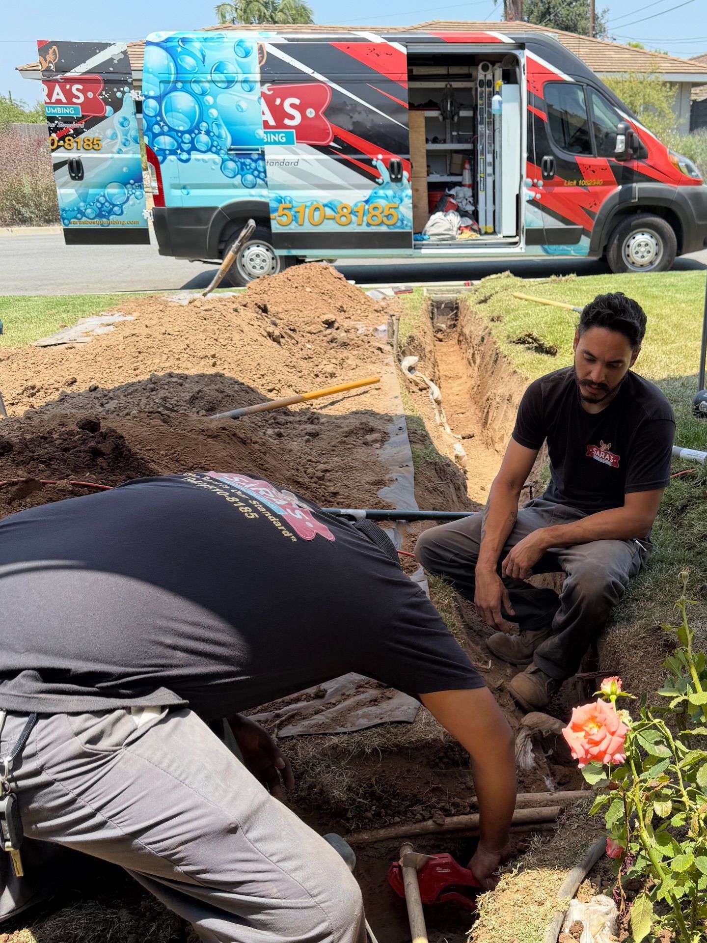 Two men in a trench working on pipes, with a van in the background. Sunny day.