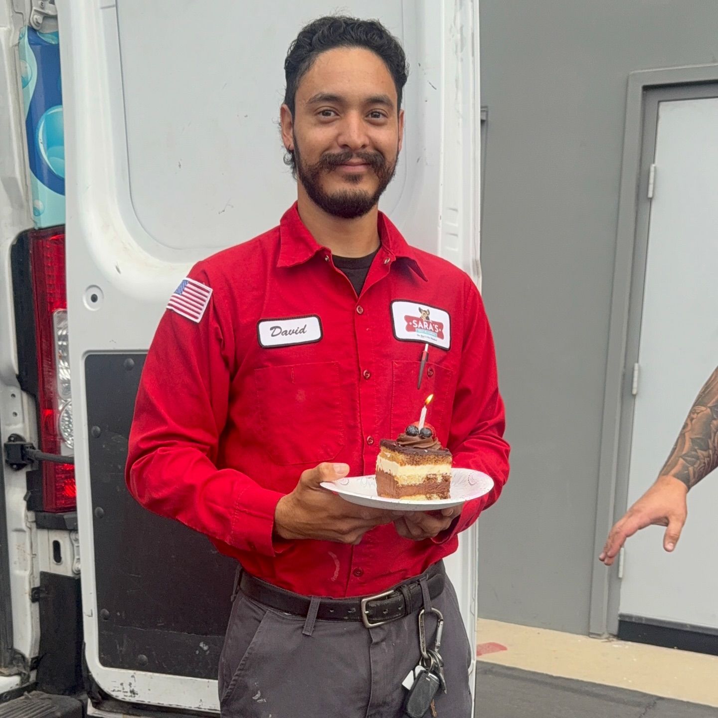 Man in red shirt holding a cake with a lit candle, standing near a van.