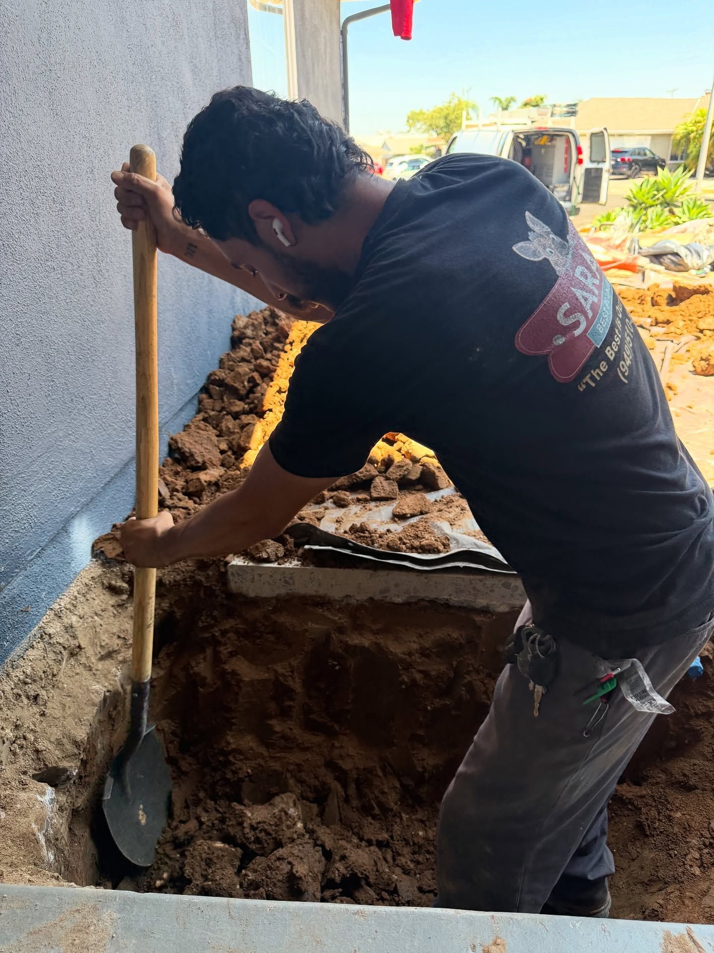 Man digging in dirt near a building's foundation with a shovel, wearing a black t-shirt in a sunny outdoor setting.