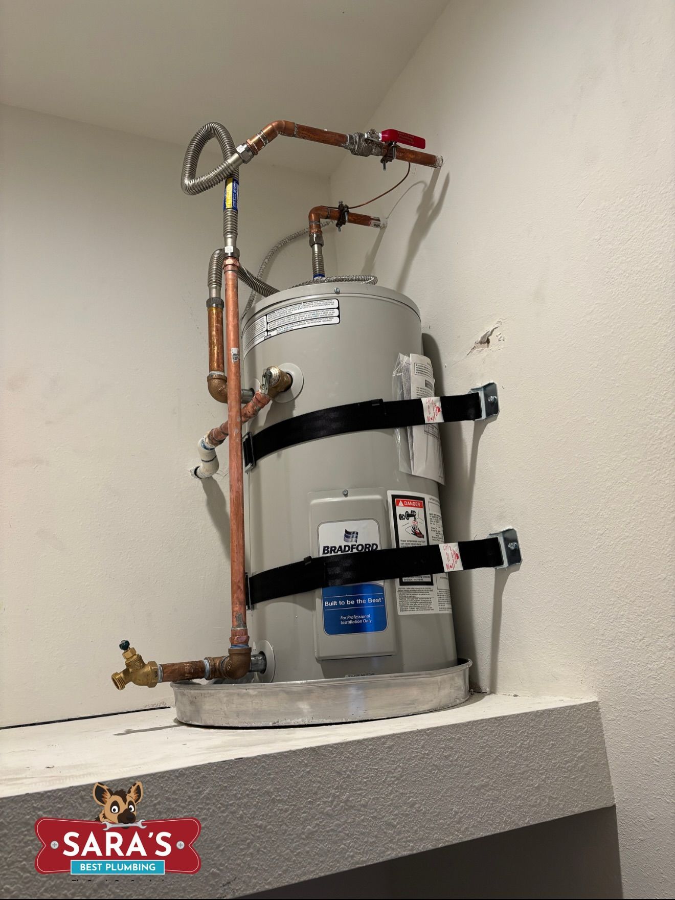 A water heater in a corner. Copper pipes, gray tank, red valve. Dark bands secure the tank.
