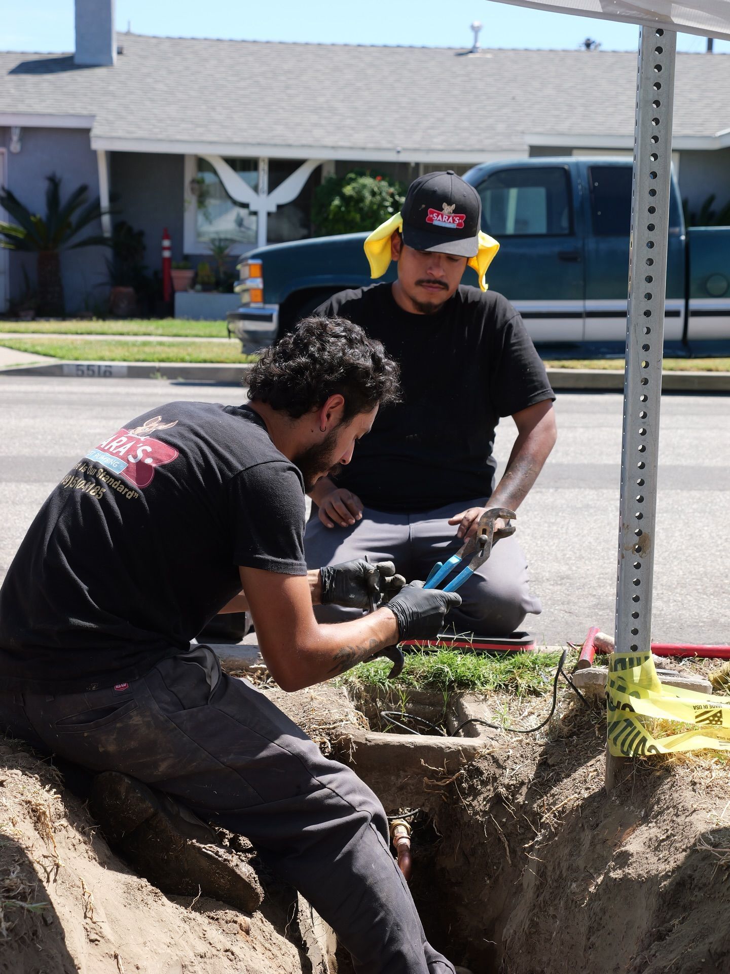Two men installing something in a roadside hole; one kneels, cutting a wire. A house and truck are in the background.