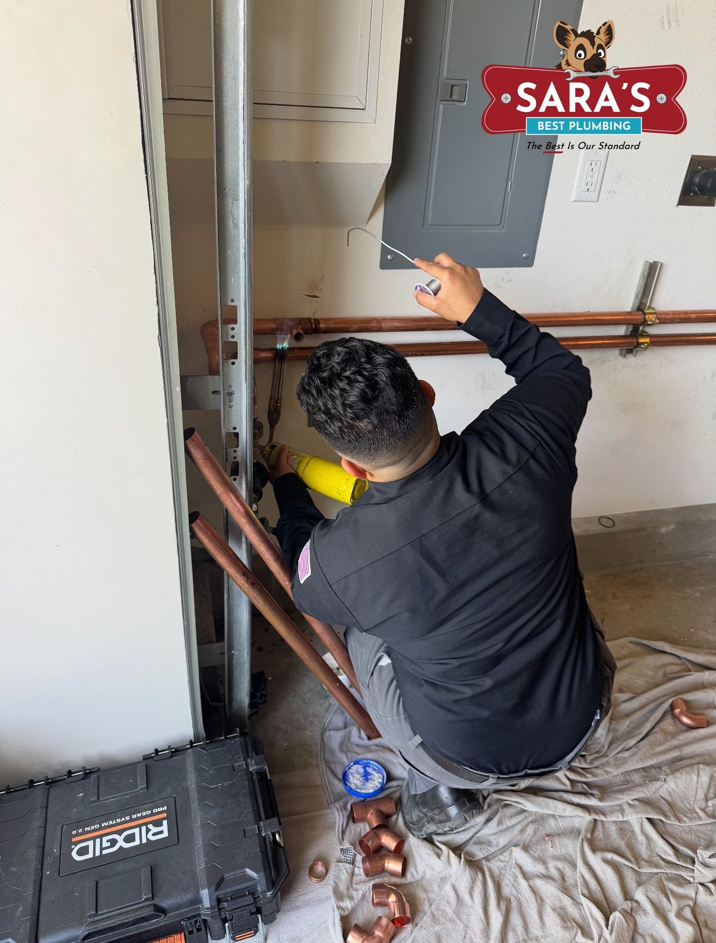 Plumber works on copper pipes near garage door. He wears black and gray, pointing at work.