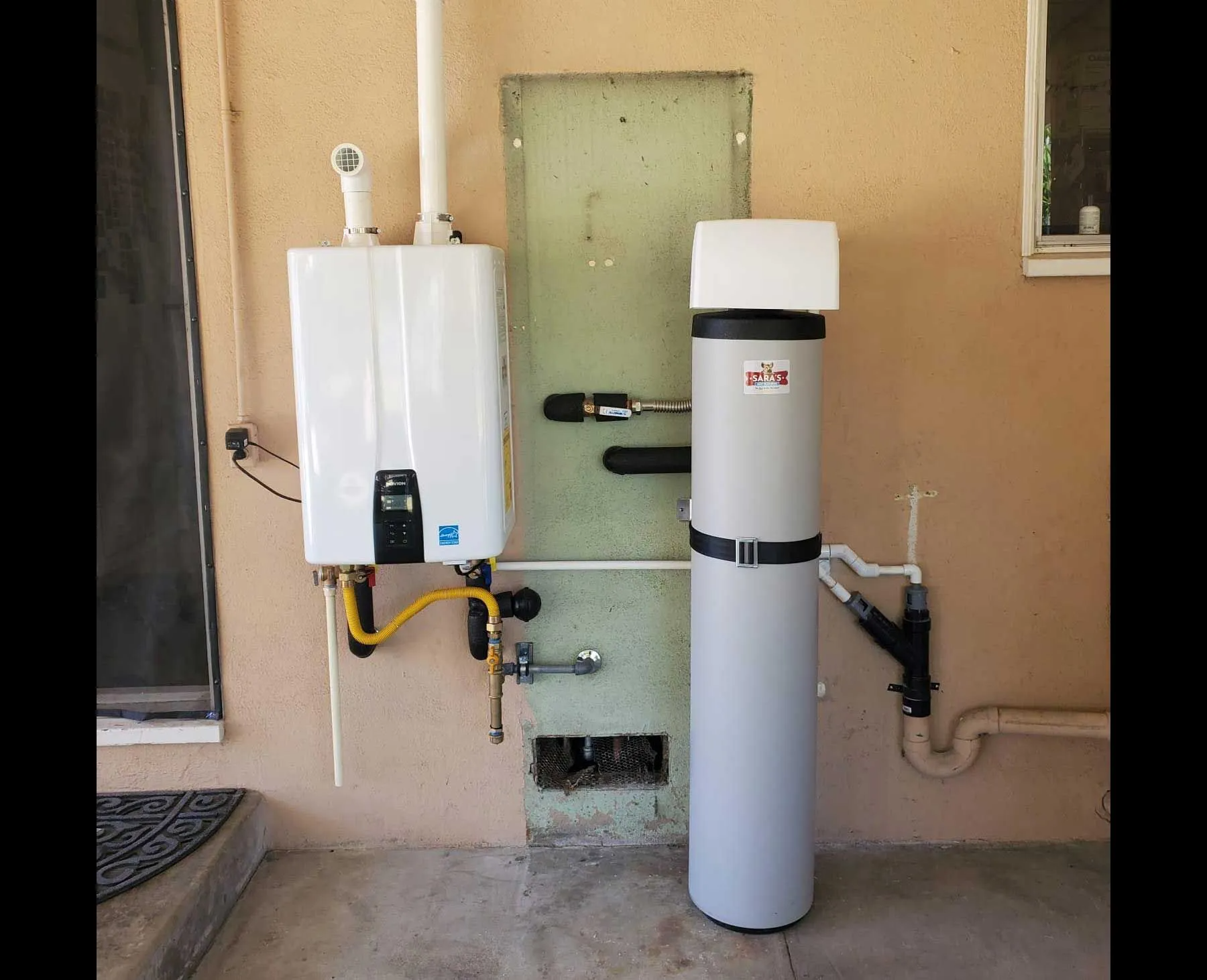 A gas water heater and water softener installed outdoors on a stucco wall next to a window.