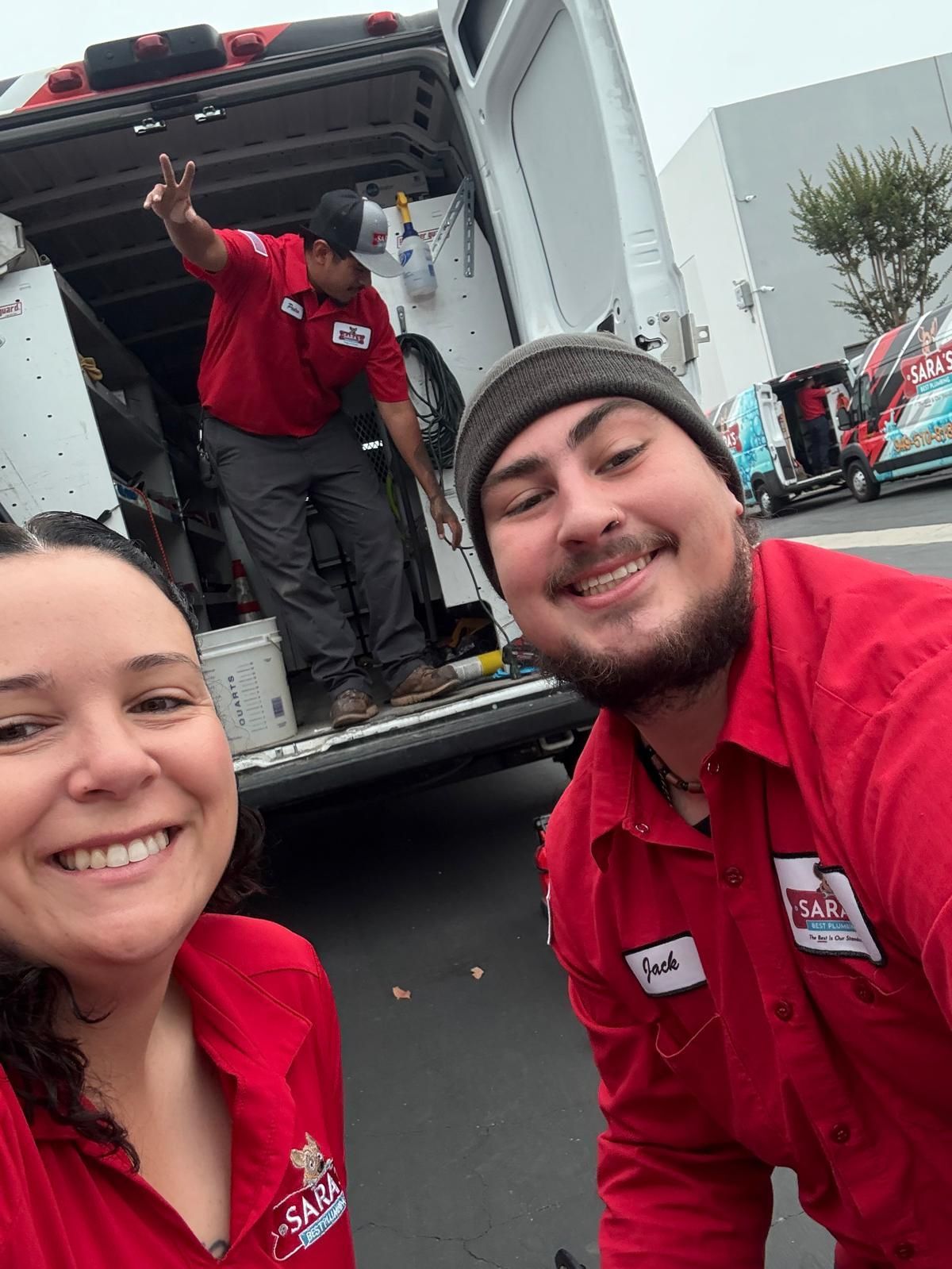 Three people in red shirts smile, with a van in the background. One points, two are near the camera.