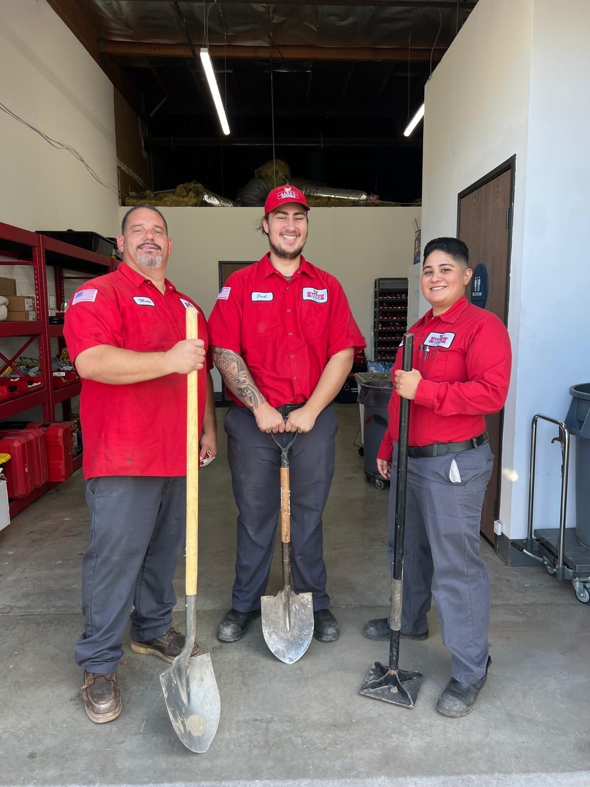 Three people in red shirts holding shovels, standing in a garage.