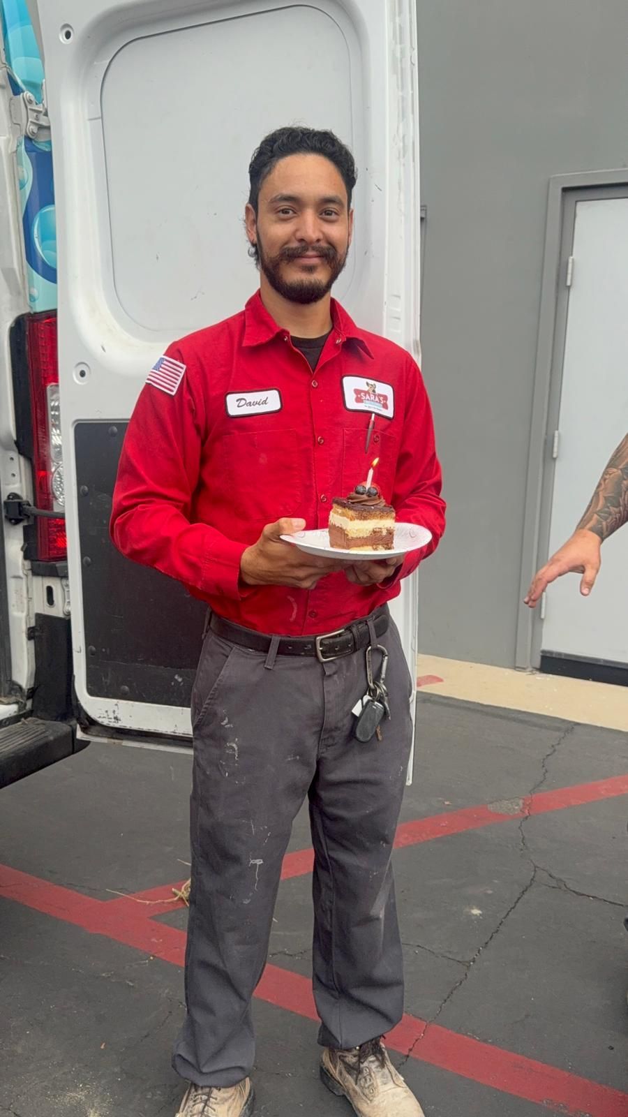 Man in red shirt, holding a small cake with a lit candle, standing outside a van.