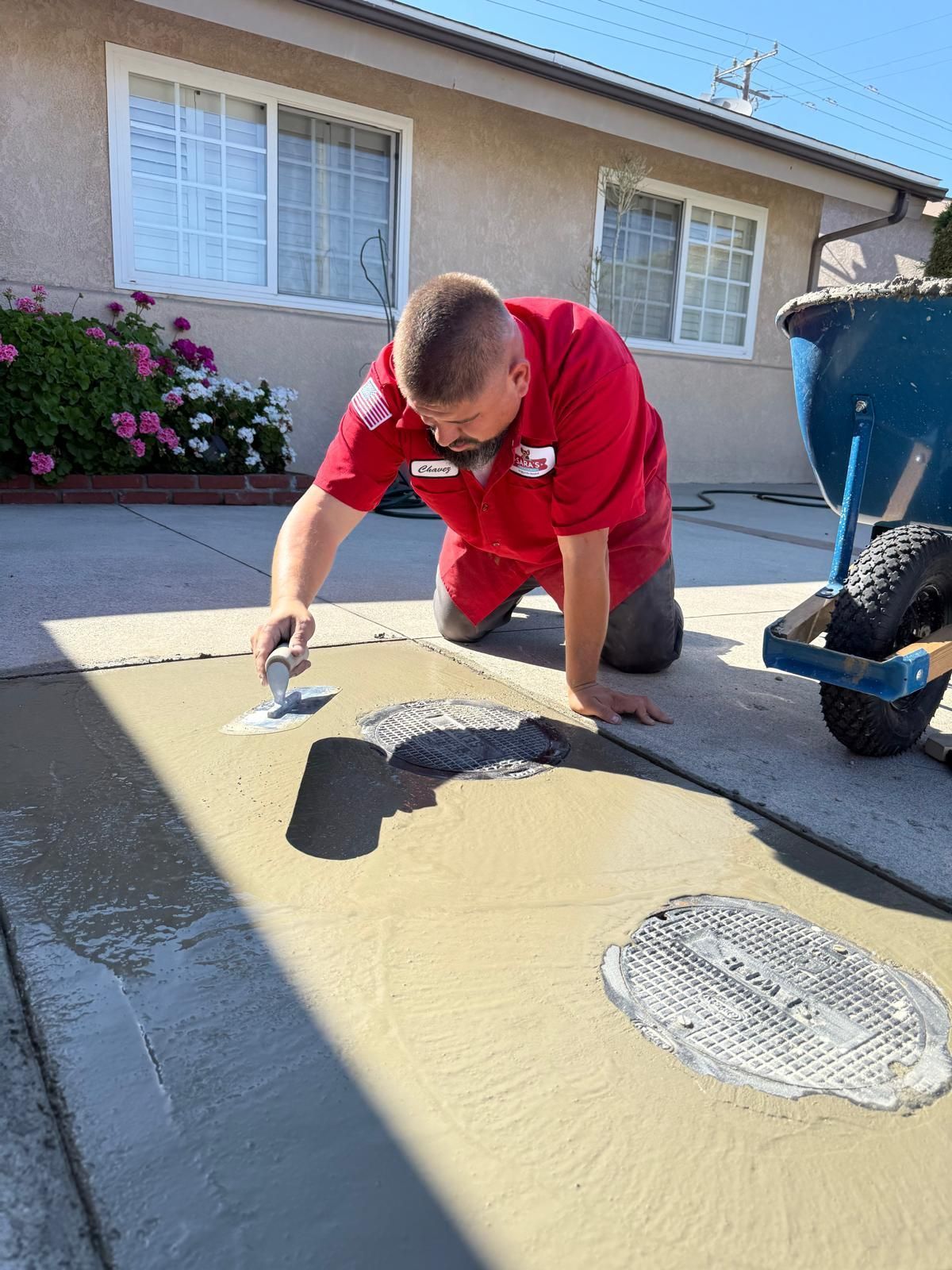 Man kneeling, smoothing cement on a sidewalk. A blue wheelbarrow and a house in the background.