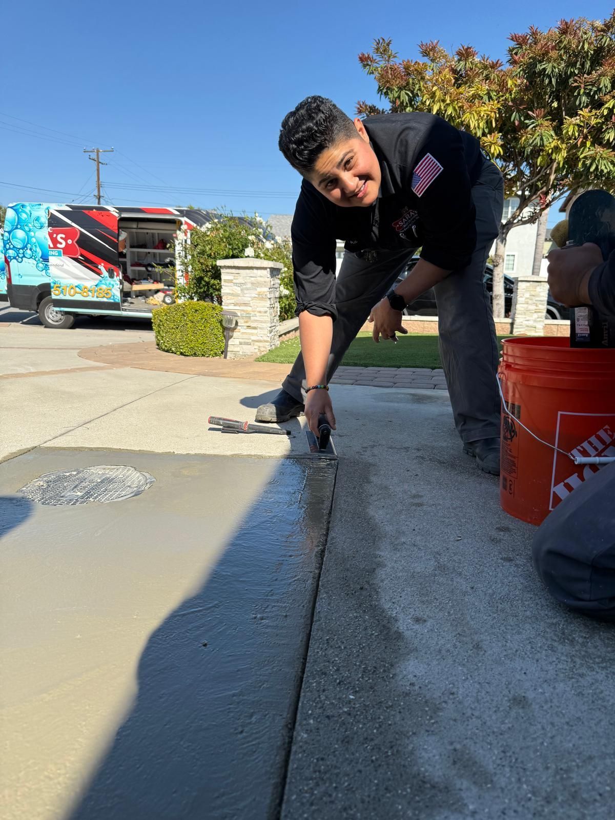Person smoothing fresh concrete on a driveway. Sunlight, blue sky, Home Depot bucket visible.