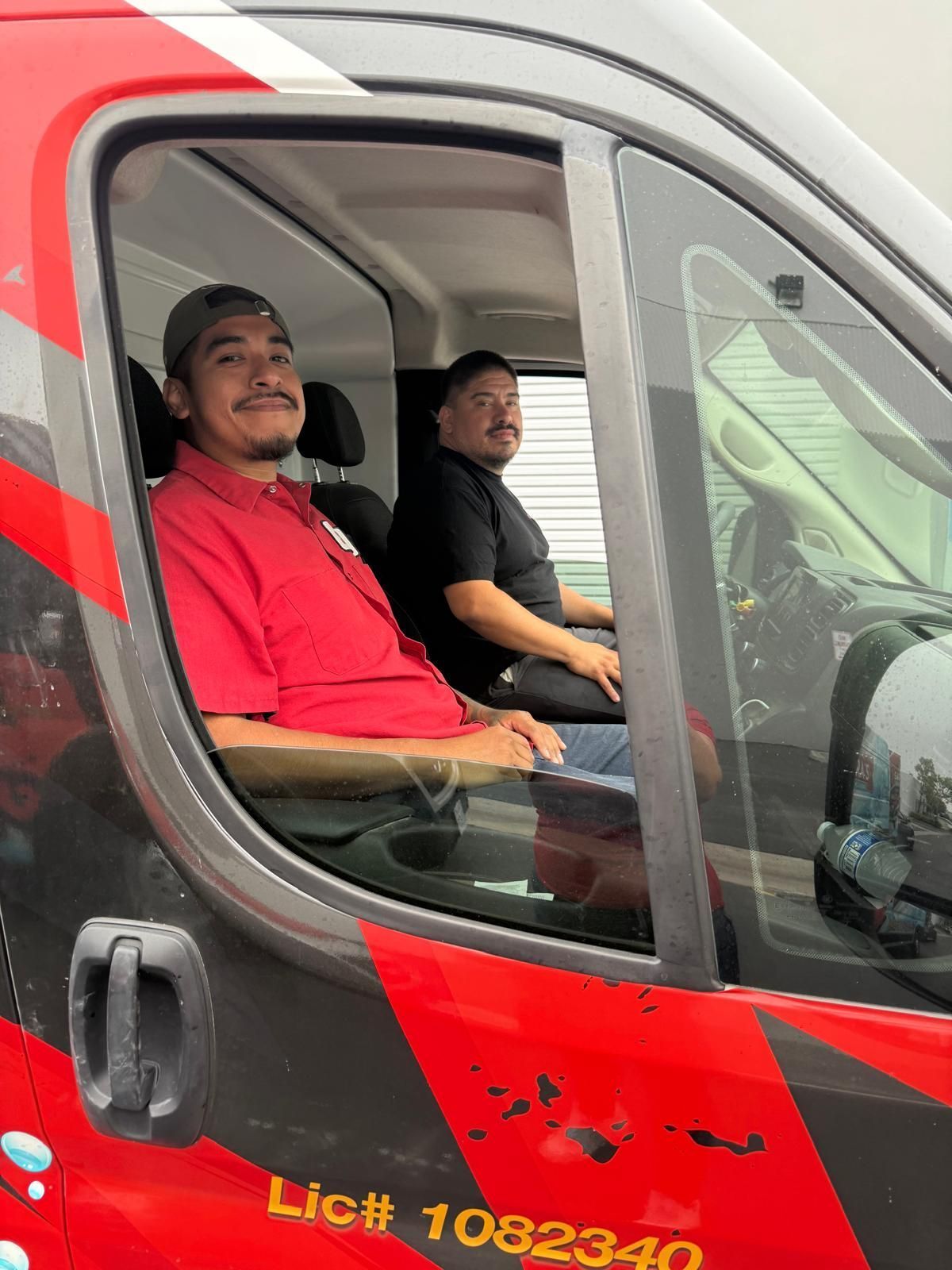 Two men in a van; one wearing red, the other black. Both look at camera. Van is red and gray.
