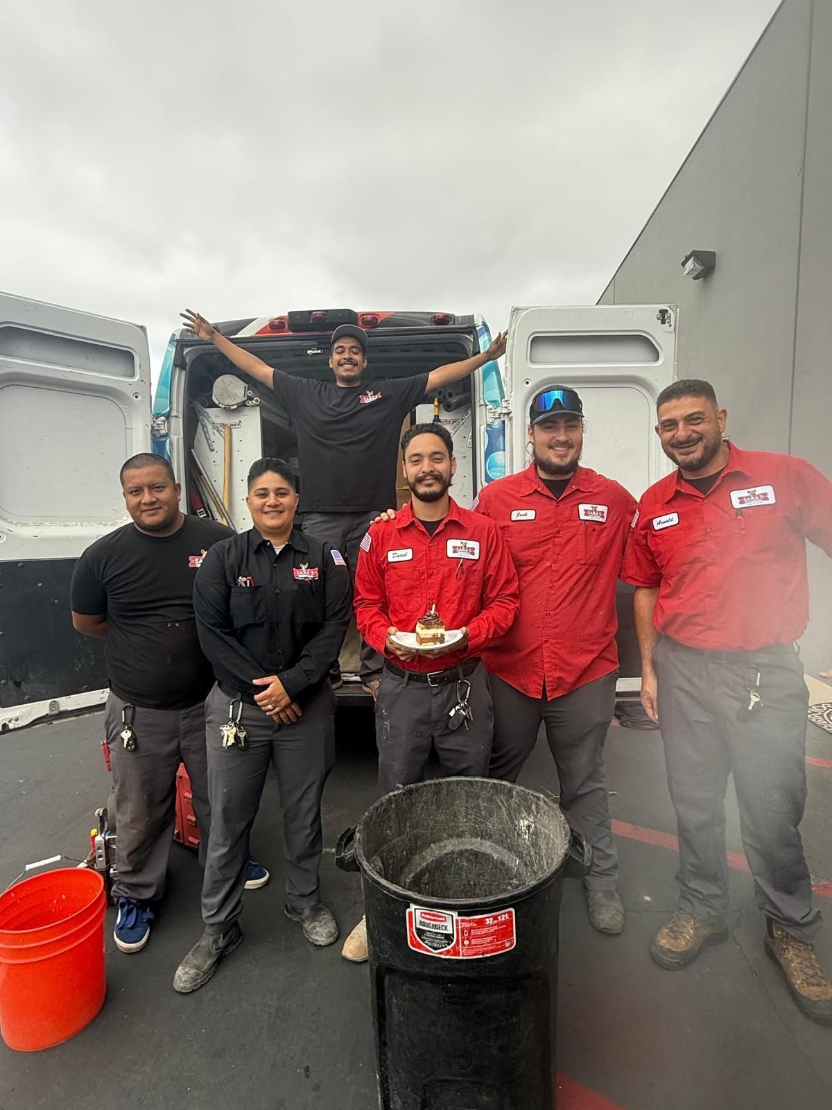 Group of technicians in work uniforms pose near a van and trash can, smiling.