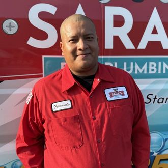 Man in red uniform smiles, standing in front of a service truck, wearing a baseball cap; company logo visible.