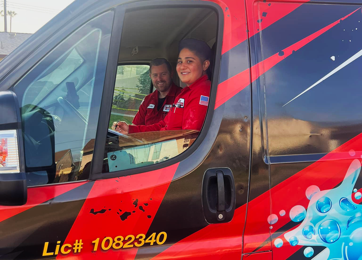 Two people in red uniforms in a service van, smiling. Van has black and red design, license number.