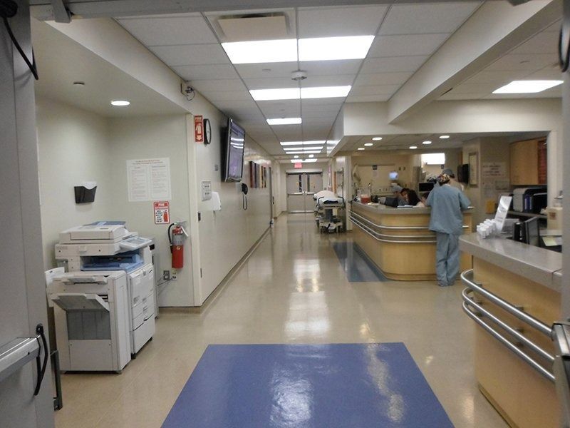 Hospital corridor with a reception desk and staff, lit by fluorescent ceiling lights.