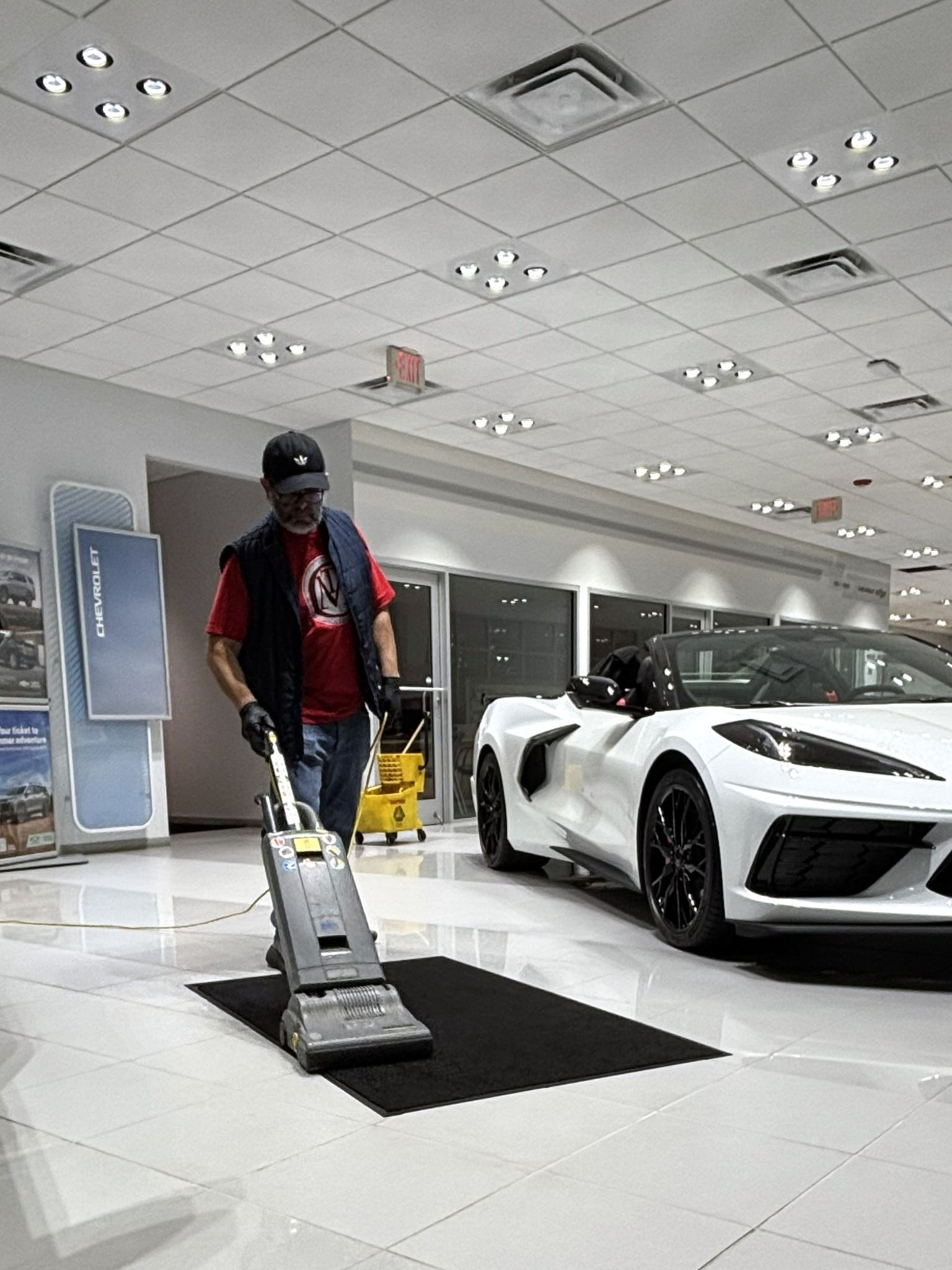 Person in a showroom vacuuming near a white sports car under bright ceiling lights.