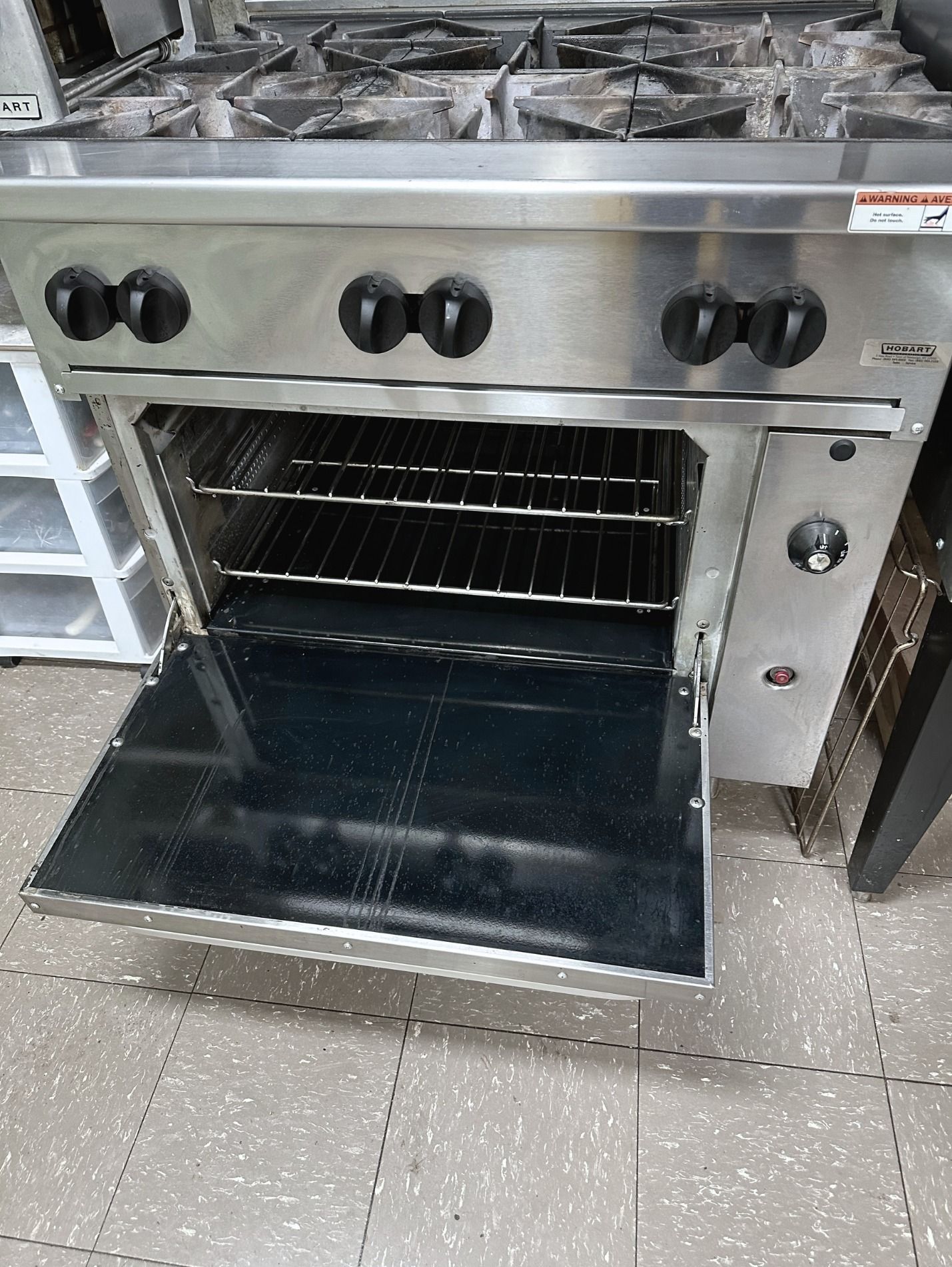 Stainless steel commercial oven with open door below stovetop in a kitchen setting