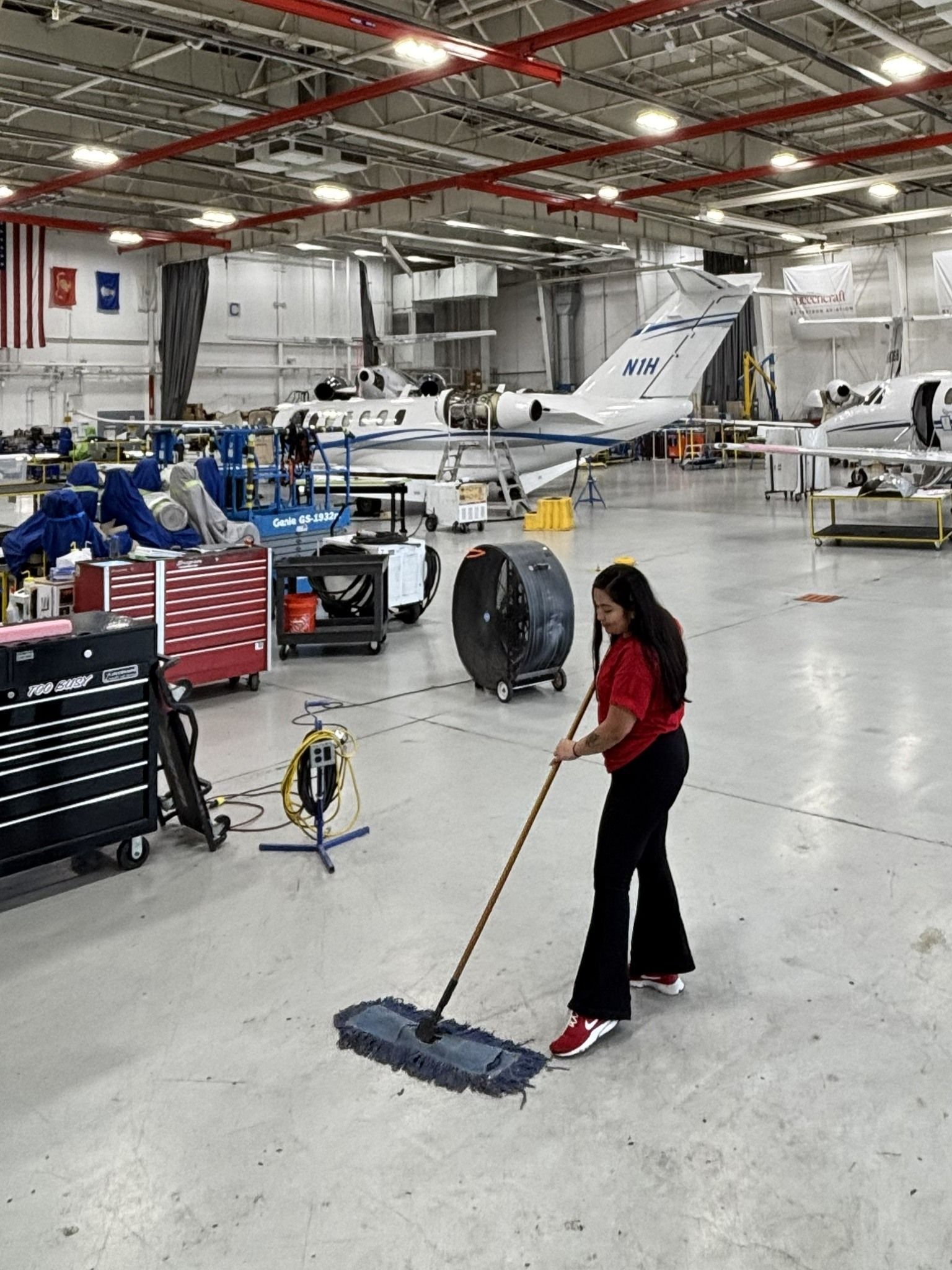 Person mopping an aircraft hangar floor with planes and equipment in the background