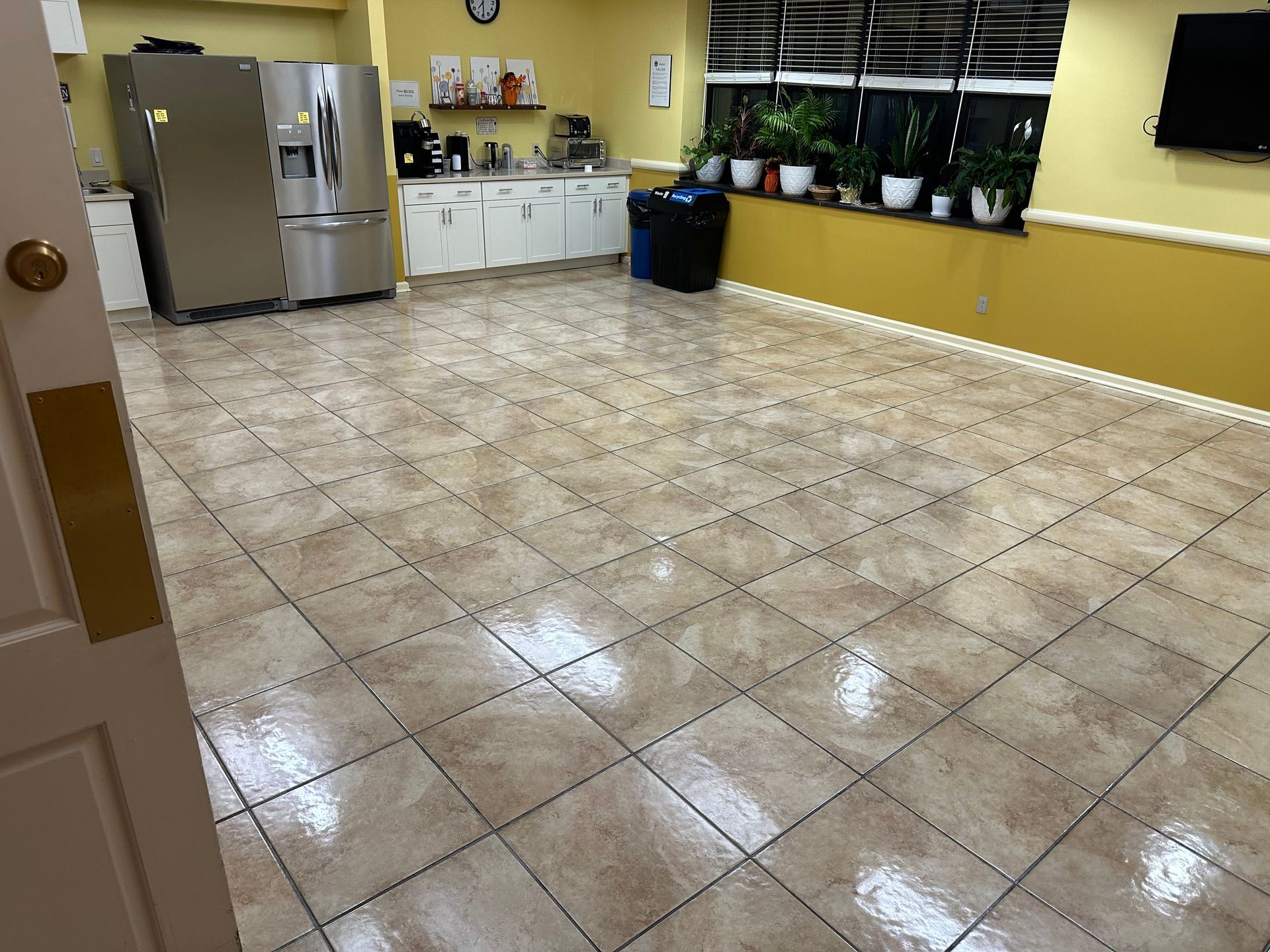 Empty kitchen with beige tiled floor, yellow walls, stainless fridge, white cabinets, and windows with potted plants