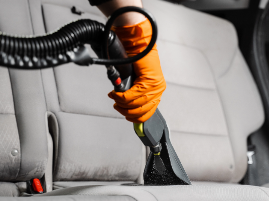 Person wearing orange glove vacuums car seat with black hose.