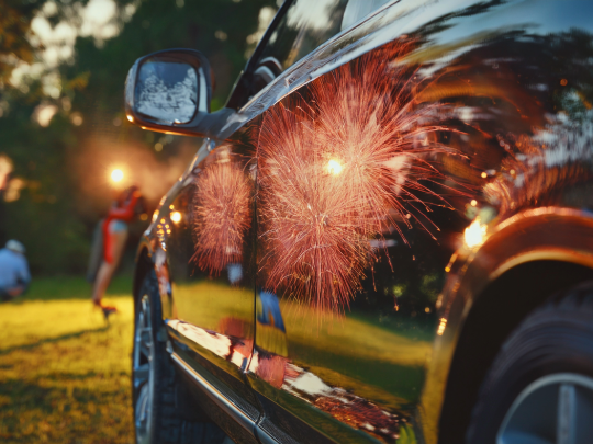 Fireworks reflected on a car's glossy black paint during an outdoor celebration.