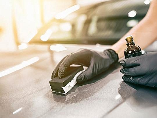 Person in black gloves applying ceramic coating to a car's surface with a sponge applicator and small bottle.