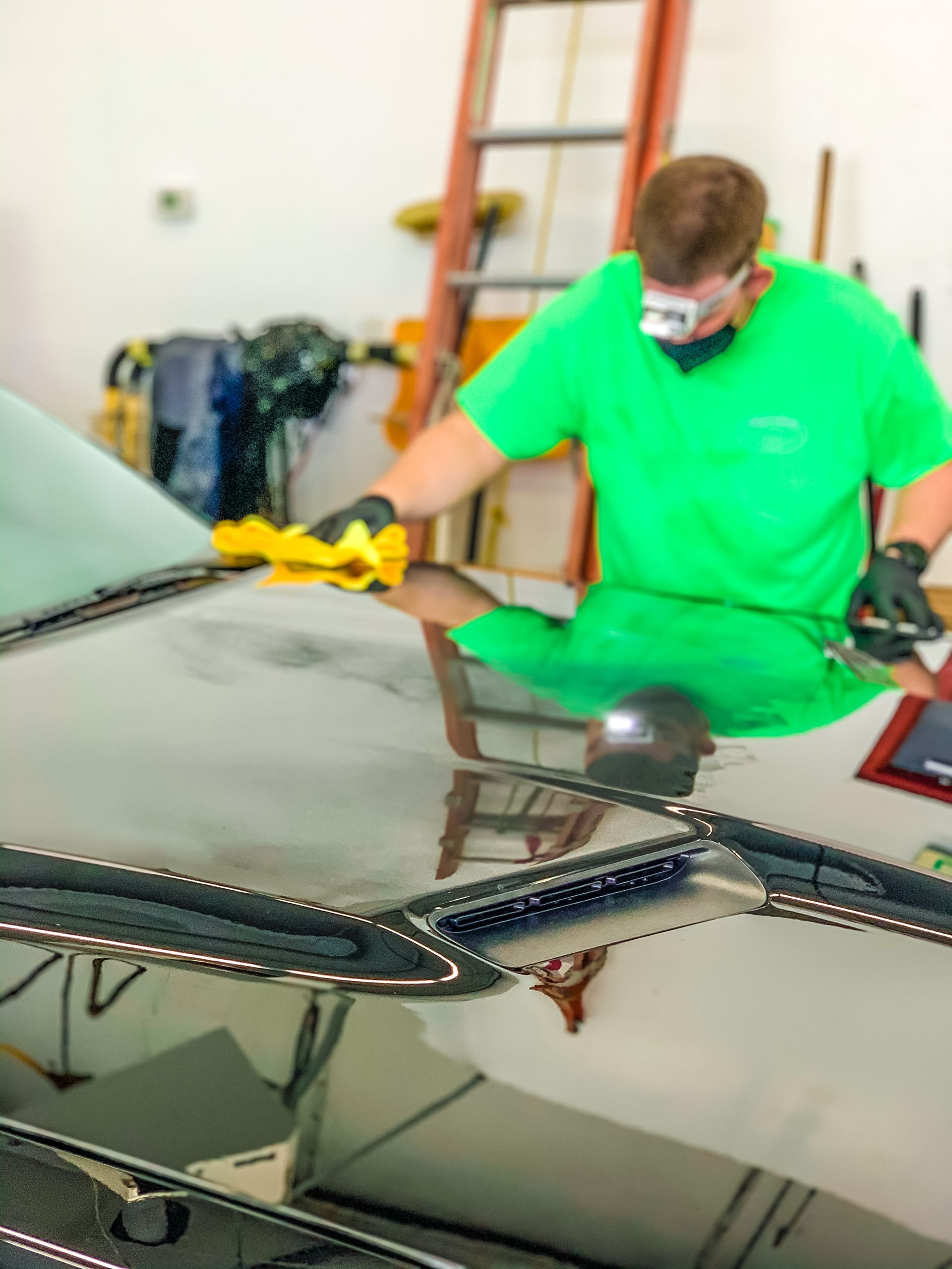 Person wearing mask and gloves polishes a car hood in a garage, using a yellow cloth.
