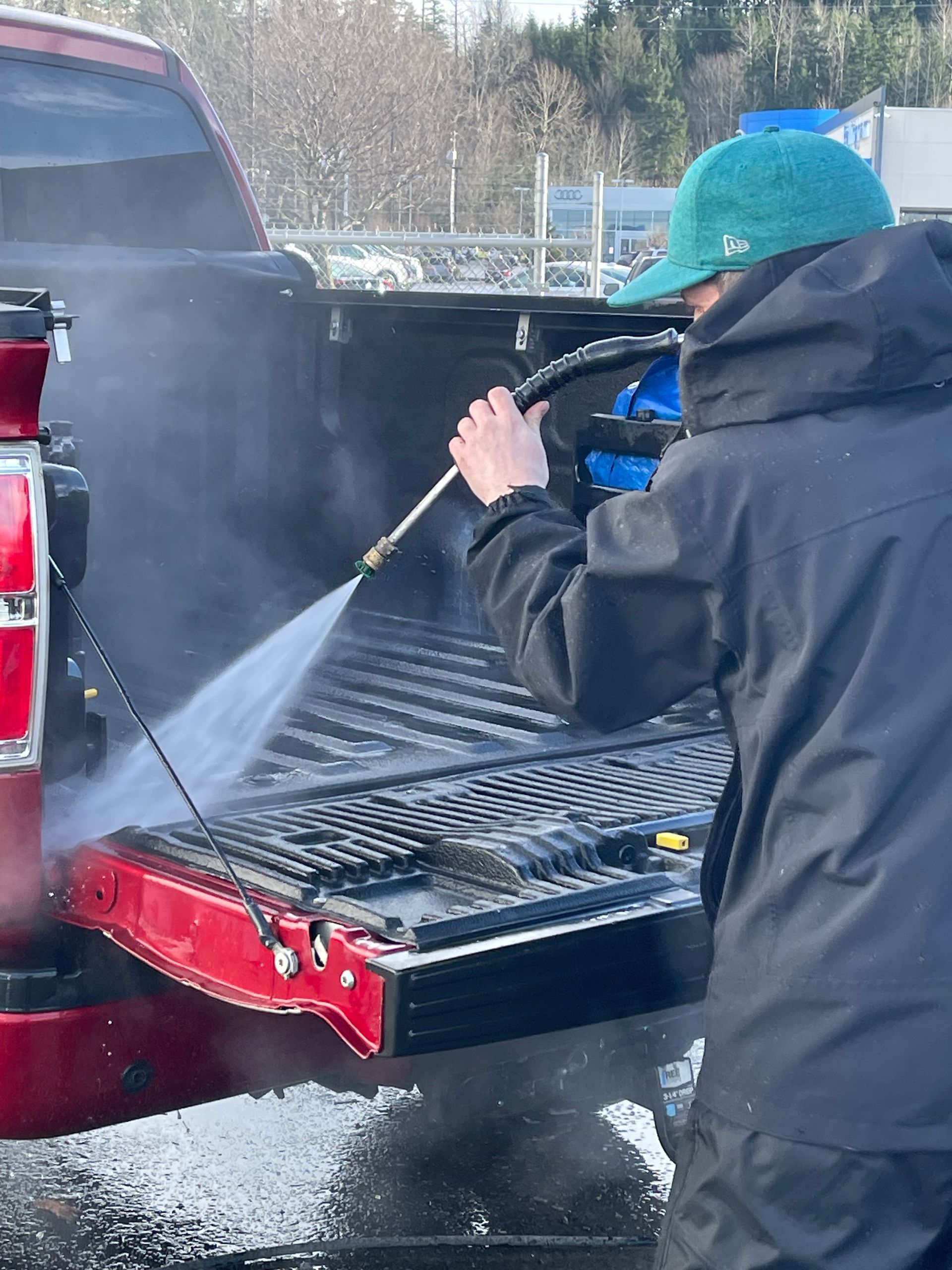 Person wearing a green hat and black jacket pressure washing the bed of a red pickup truck.