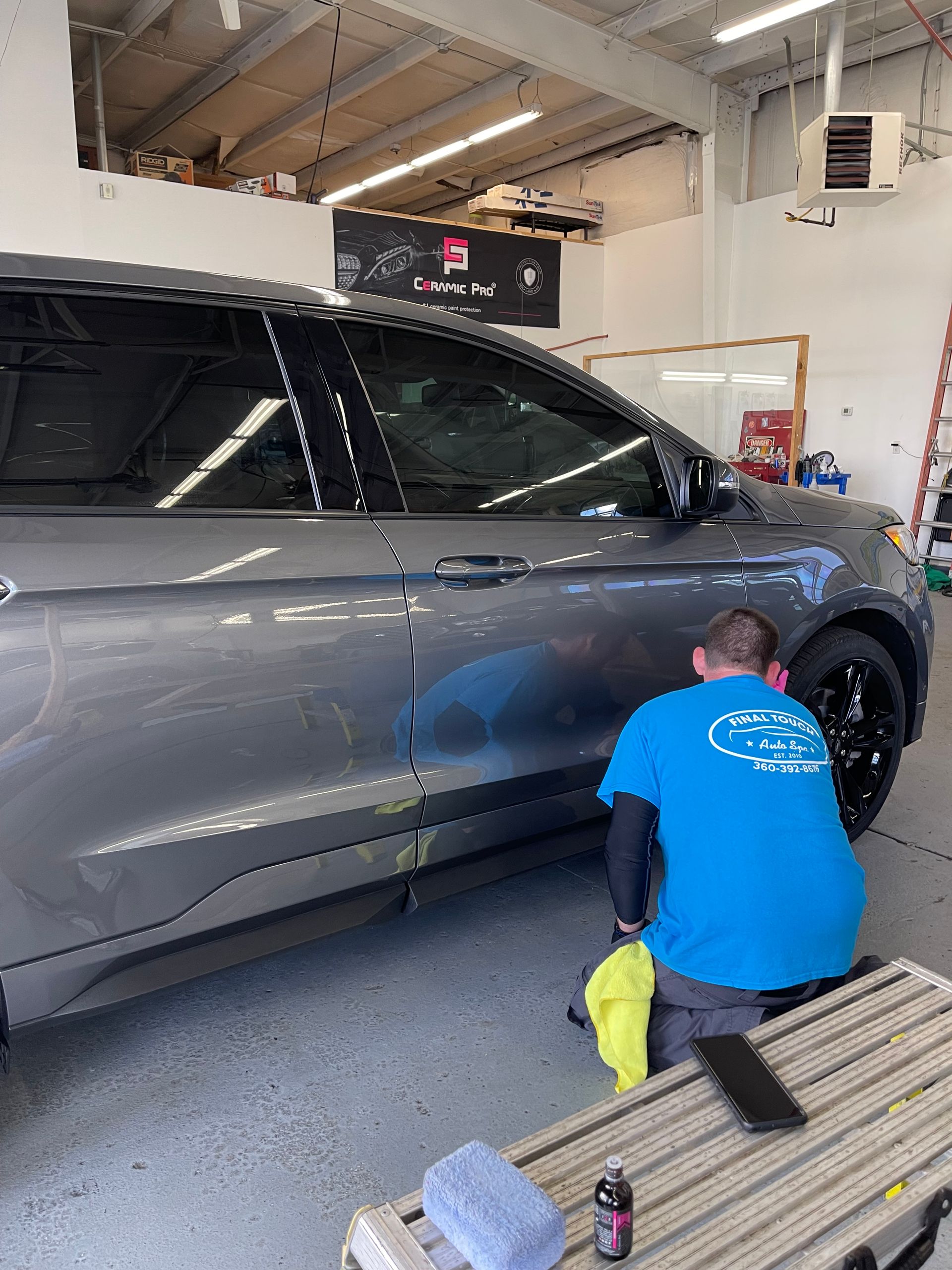 Man polishing a gray car in a shop. He wears a blue shirt and kneels beside it.