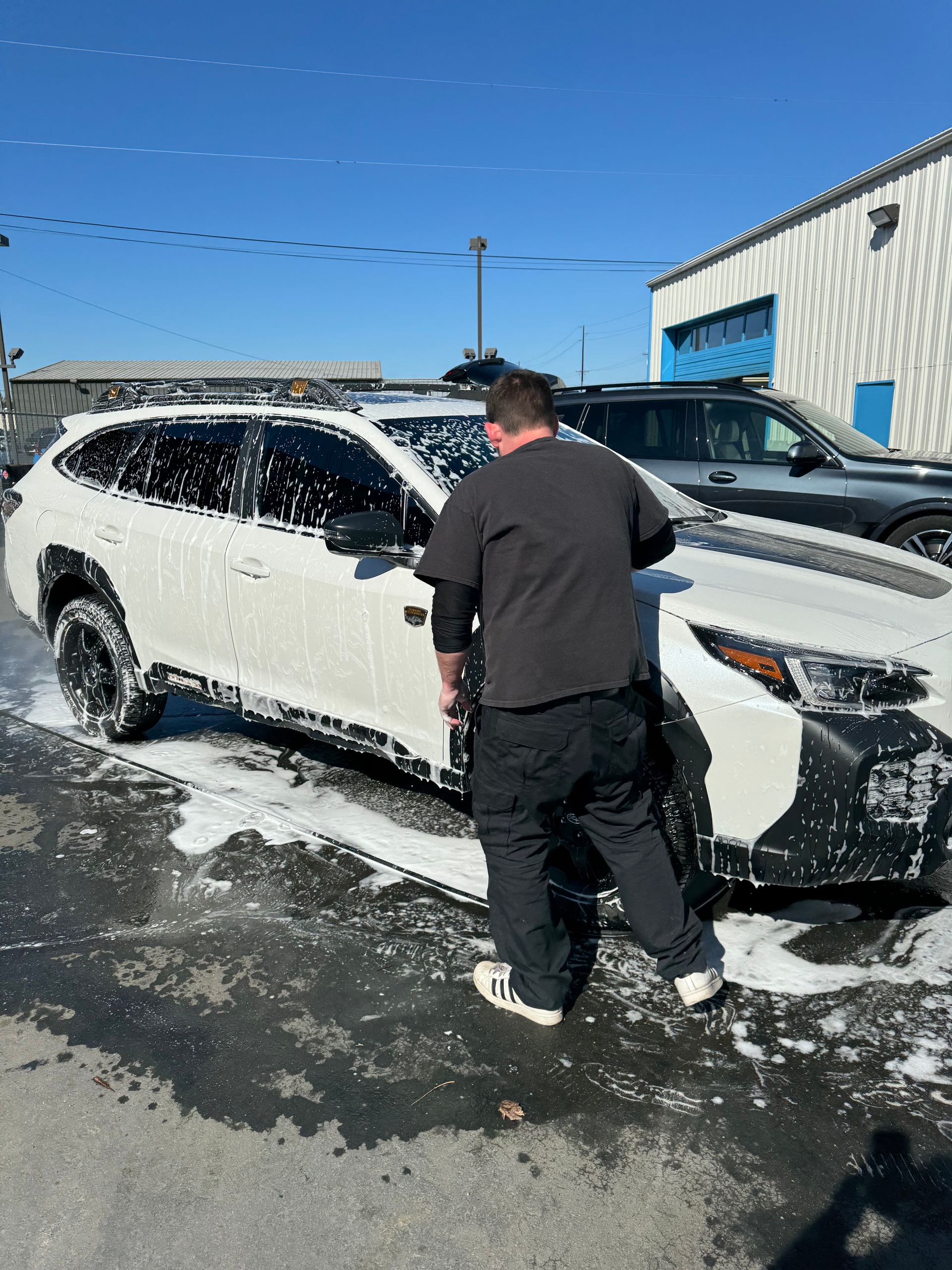 Person washing a white car with black accents outdoors in front of a building.