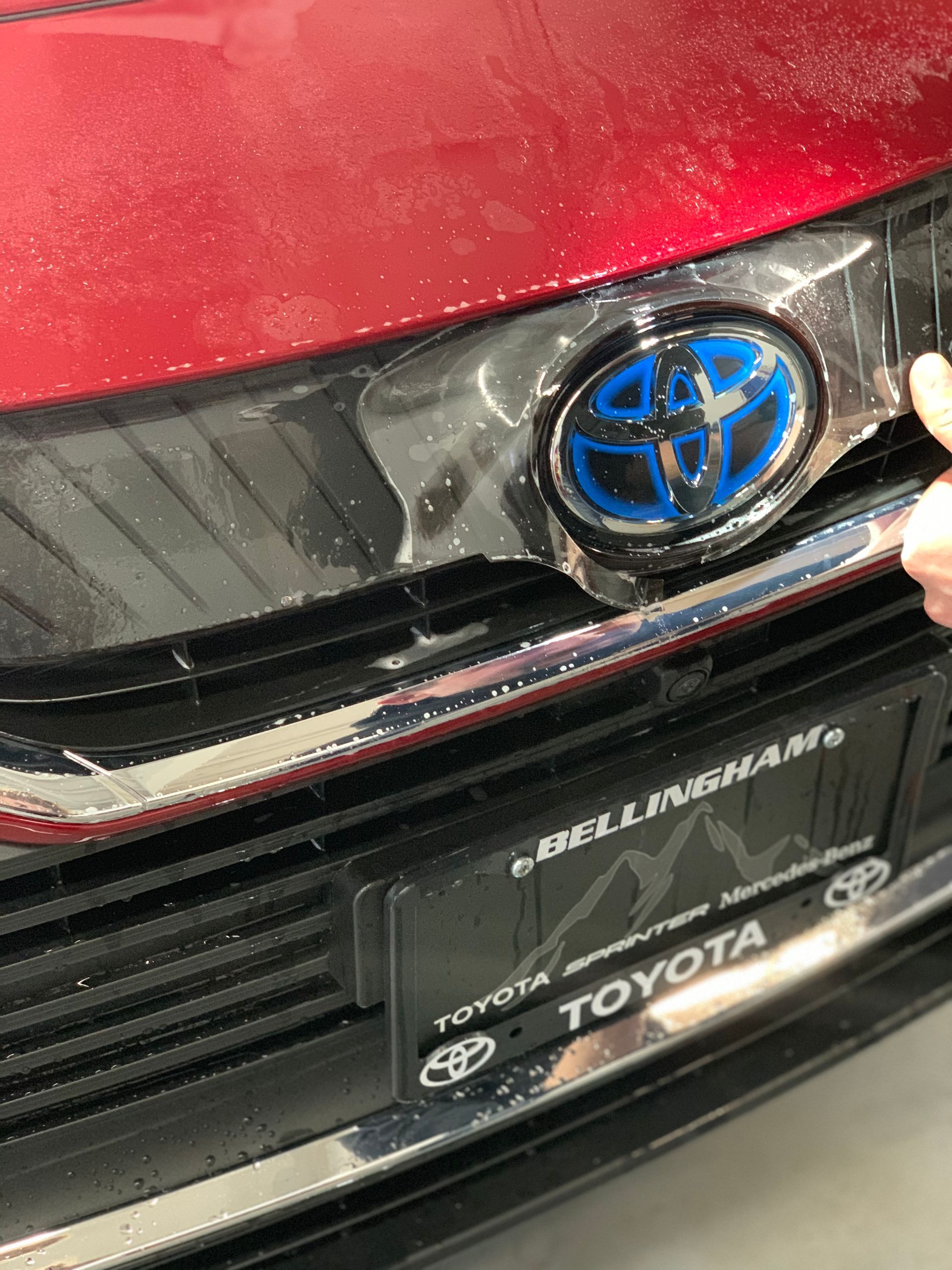 Red Toyota car with clear protective film on the front grille. Hand peels film near blue Toyota emblem.