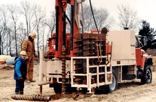 Drilling rig on a truck with two people, brown and blue clothing, on a field.