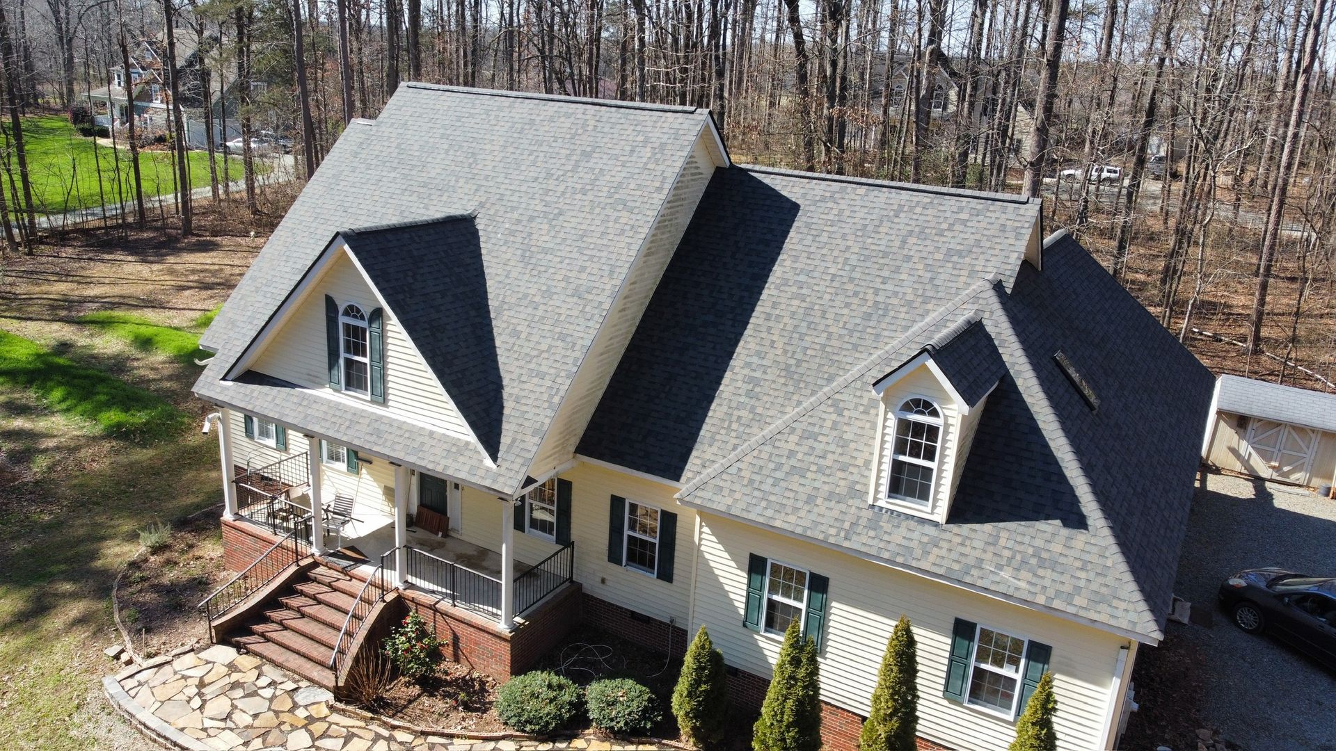 Aerial view of a gray house with a steep roof and front porch, surrounded by trees and a yard.