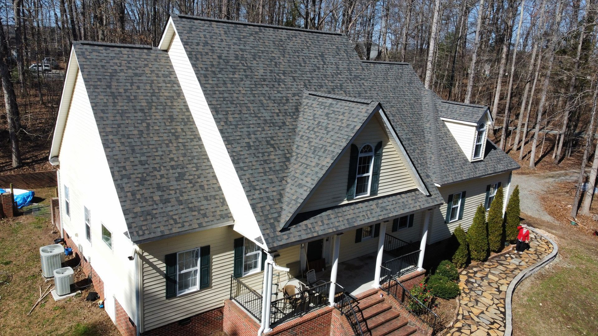 Aerial view of a white house with a dark gray roof, front porch, and landscaped yard surrounded by trees.