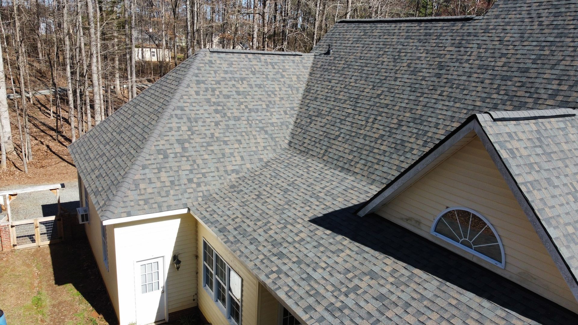 Roof of a beige house with gray shingles beside a wooded area on a sunny day
