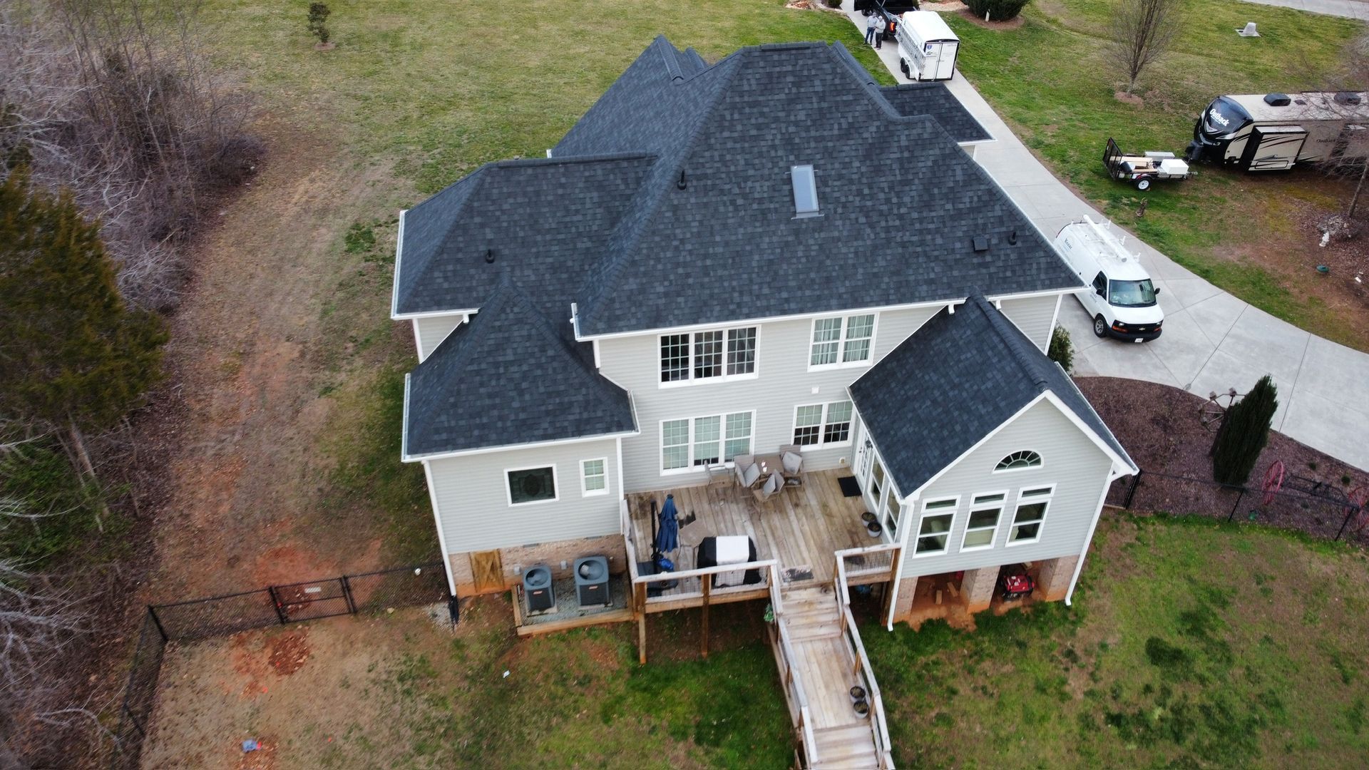 Aerial view of a large gray two-story house with a wooden deck and stairway, surrounded by trees and yard.