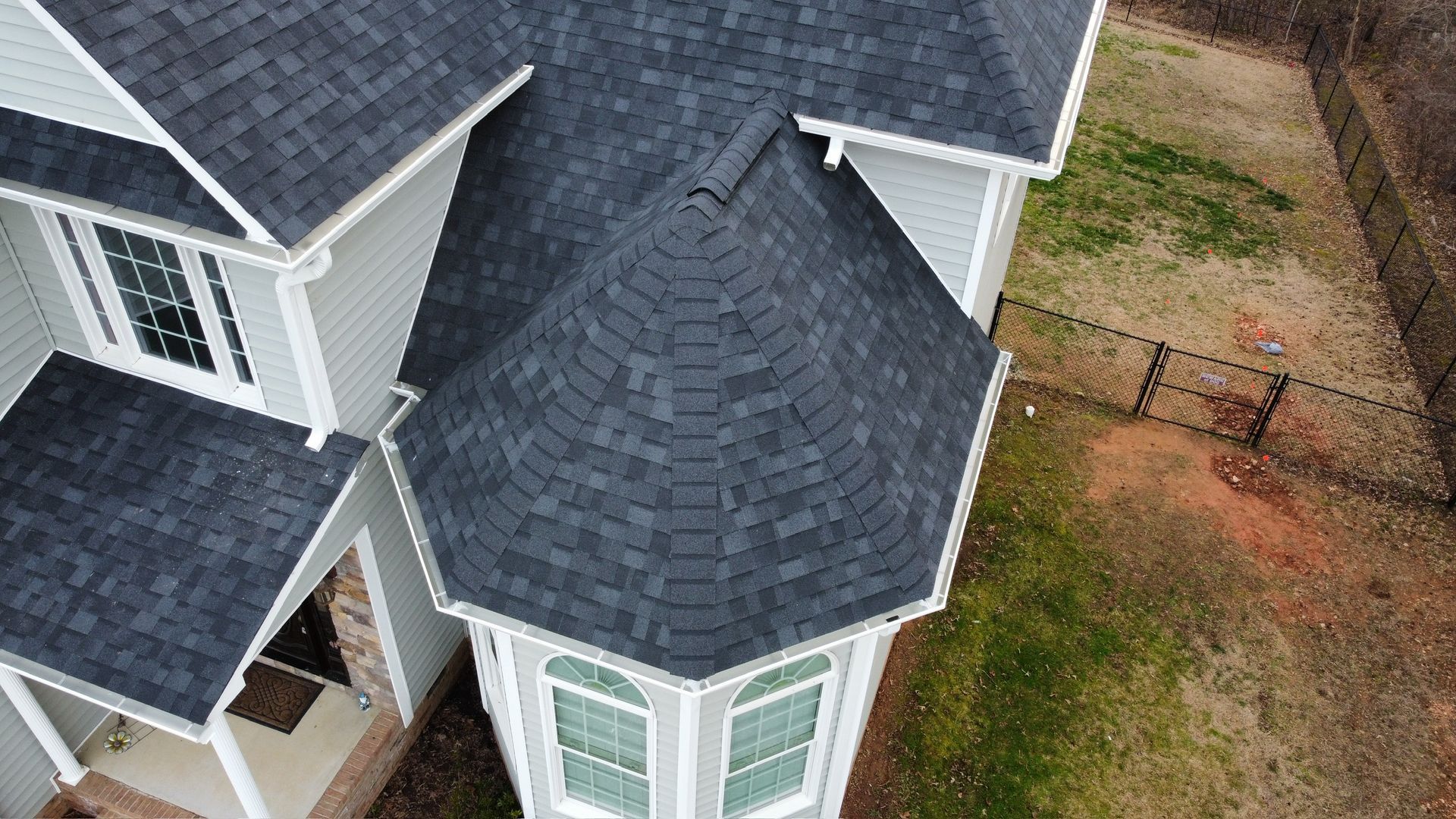 Aerial view of a house with dark gray shingle roof and white siding beside a dirt yard