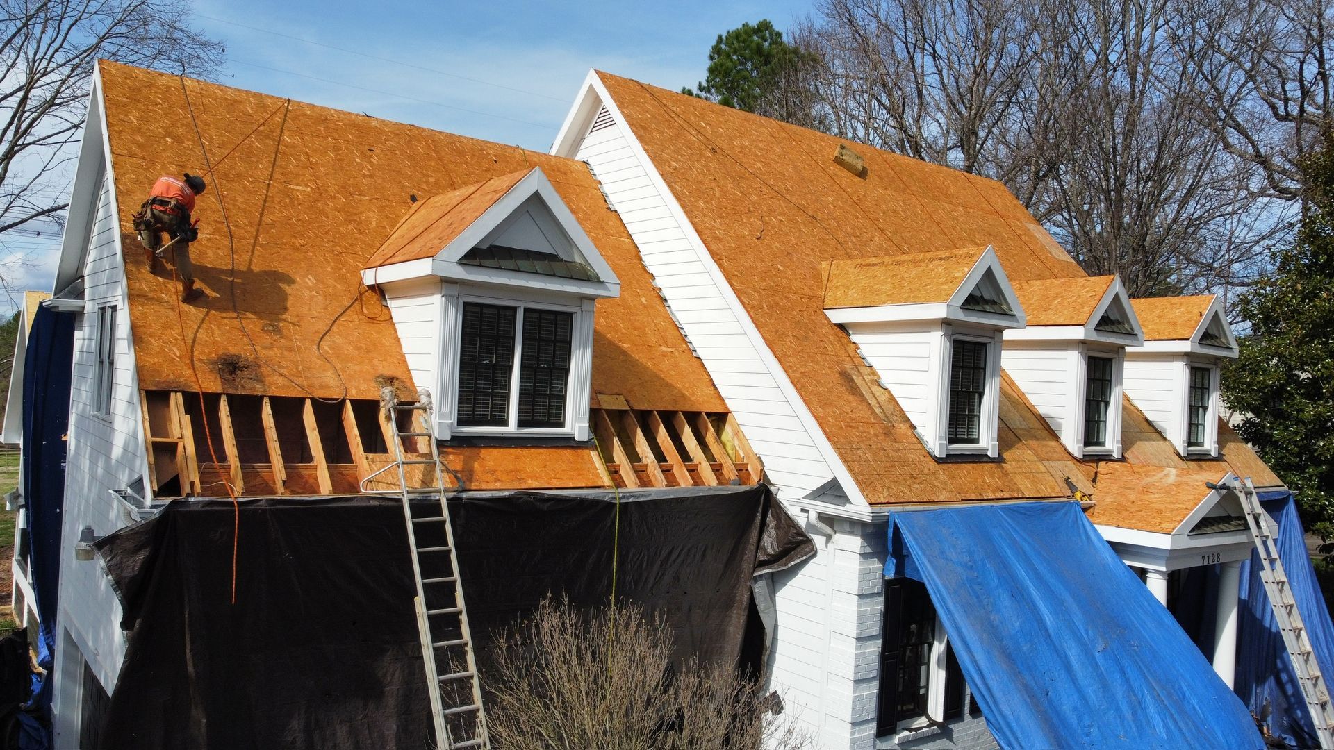 Workers on a steep orange roof, with ladders and blue tarp covering part of the house