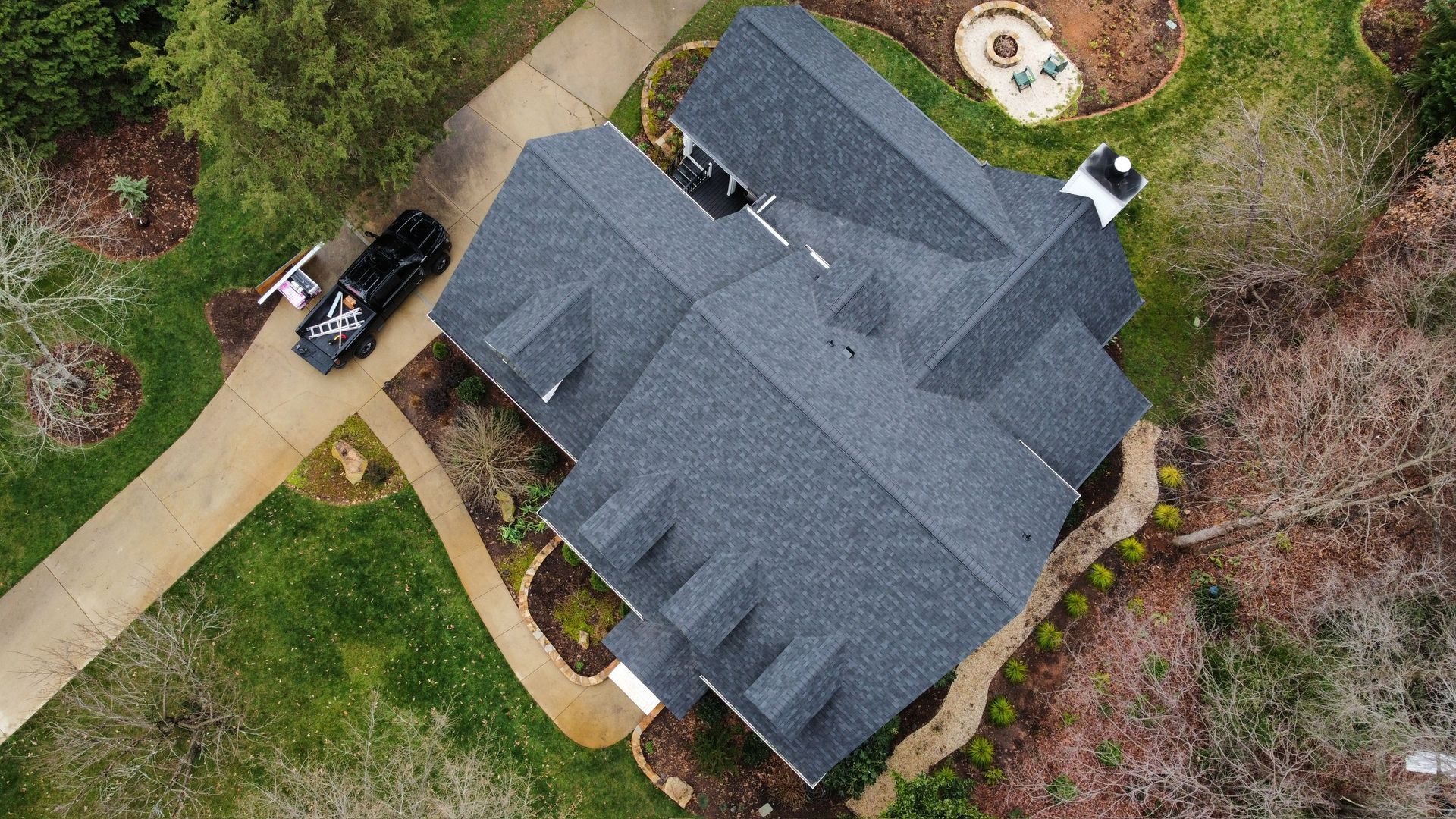 Aerial view of a gray-roofed house with a driveway, parked car, and surrounding trees and lawn.