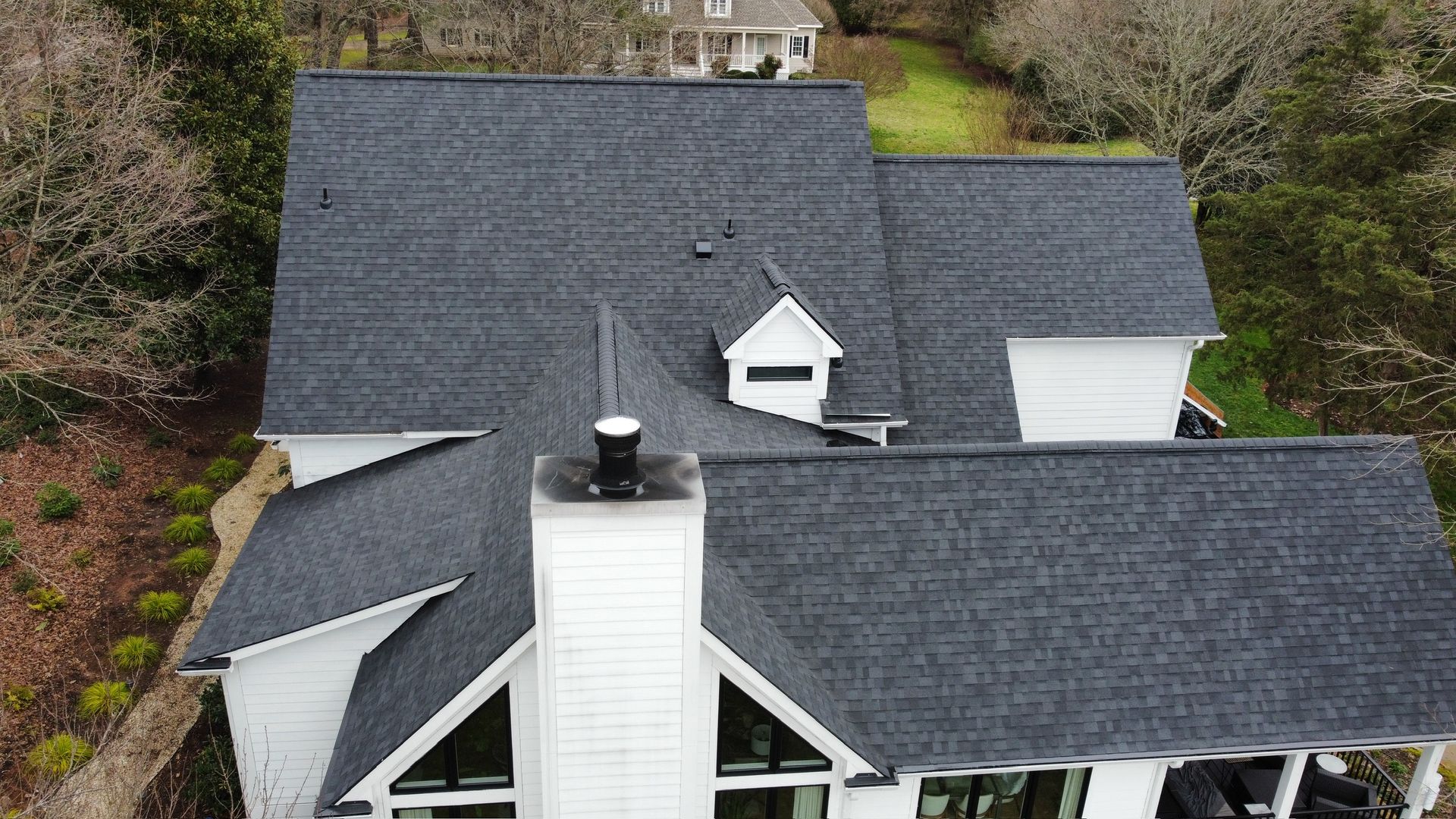 Aerial view of a house with dark gray shingled roof and white siding surrounded by trees