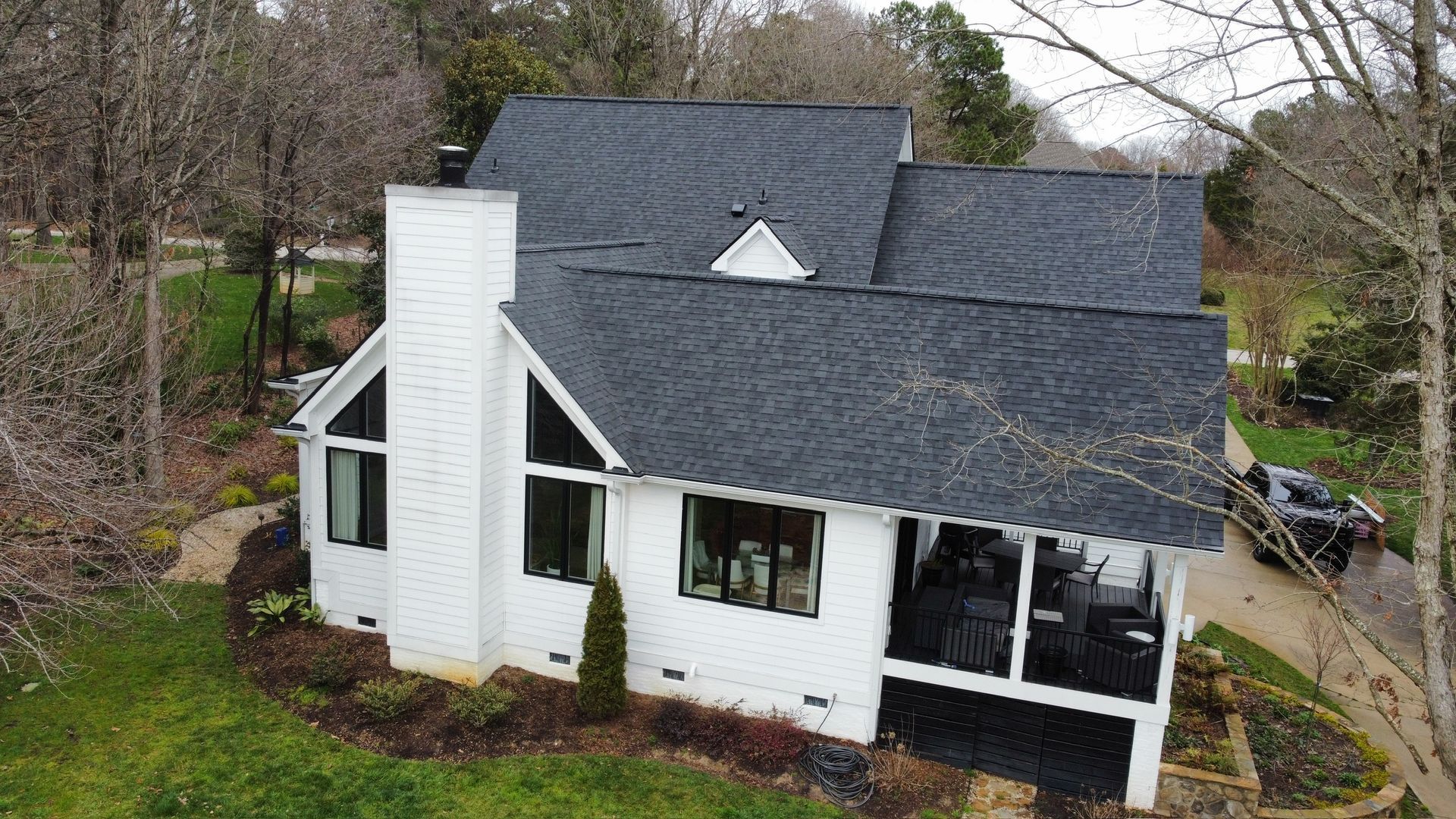 Aerial view of a house with dark gray shingle roofs and white siding, surrounded by trees.