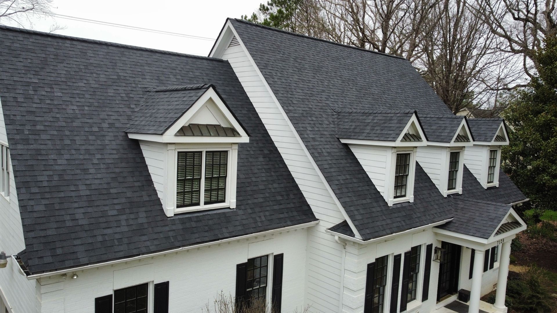 White house with dark gray shingles and multiple dormer windows, viewed from above on a cloudy day