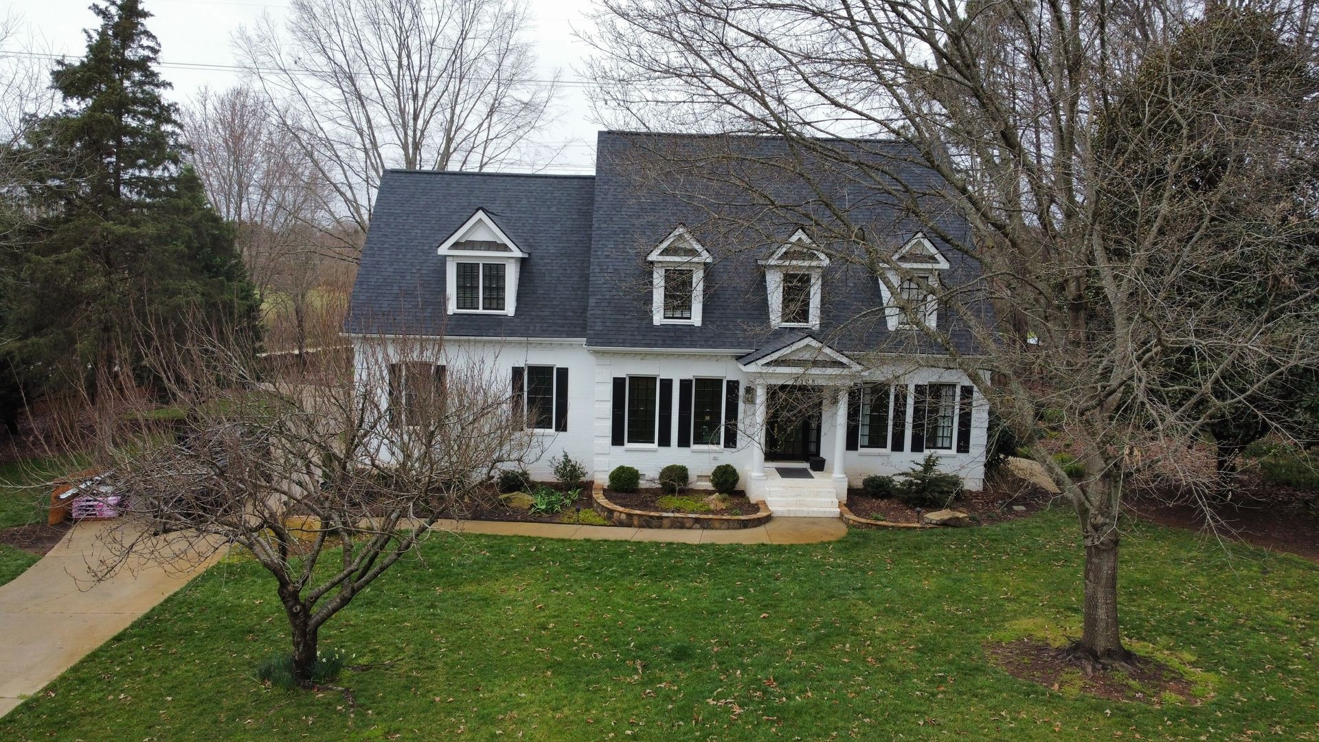 Two-story gray house with white trim and dormer windows, front yard and trees in overcast weather