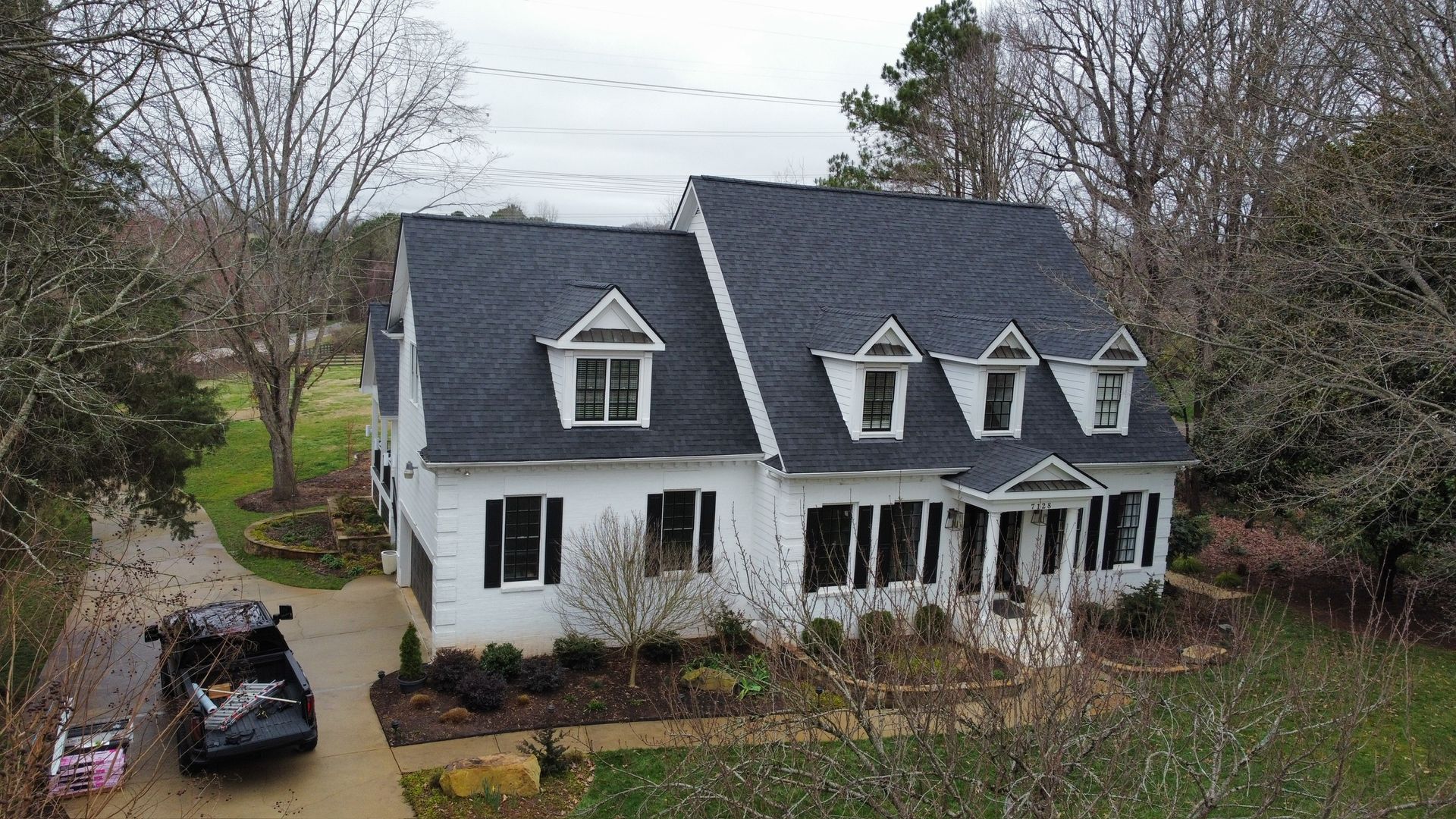 White two-story house with dark roof and front porch beside a driveway and trees